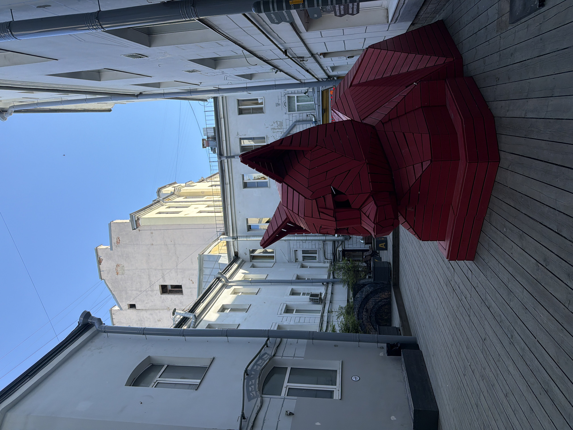 This is a street-level, medium-shot photograph taken on a clear day in Saint Petersburg, Russia. The scene is a courtyard with buildings on all sides, paved with wooden planks. In the foreground and center of the image is a large, red, geometric sculpture of a cat. The sculpture is made of segmented wooden pieces and is quite abstract in its design. The cat sculpture is sitting down, facing left in the frame.
The buildings surrounding the courtyard are multi-story structures, appearing to be a mix of residential and commercial. The building to the left is light gray with several windows and downspouts. The building to the right is white with a more modern facade, also featuring windows and downspouts, and has a sign with Cyrillic text visible. In the background, more buildings are visible, some of which appear older and show signs of wear. The sky is a clear, bright blue.
There are no people visible in the image. Some planters with small evergreen trees are placed in the courtyard, along with a dark-colored bench to the left. The overall impression is a somewhat enclosed, urban space that has been enlivened by the presence of the large red cat sculpture.
