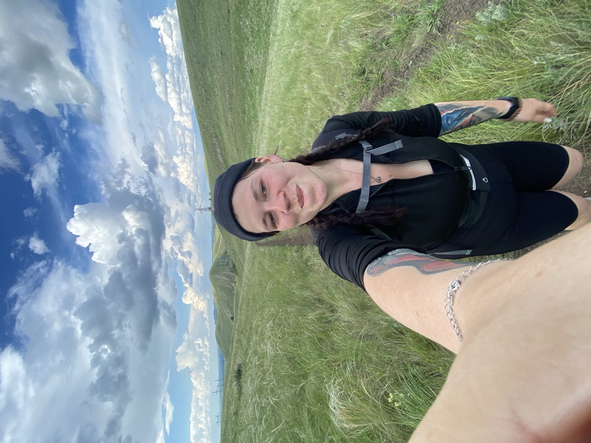 A young woman with dark braided hair, wearing a black beanie, black zip-up jacket, and black shorts, is taking a selfie outdoors. She has tattoos on her arms and is wearing a silver bracelet. The woman is standing on a dirt path in a grassy, hilly landscape. The sky is blue with fluffy white clouds. In the background, there are more rolling green hills and some power lines. The overall impression is of a scenic hike in a rural, natural environment, possibly near Verkhneozernoe, Russia.