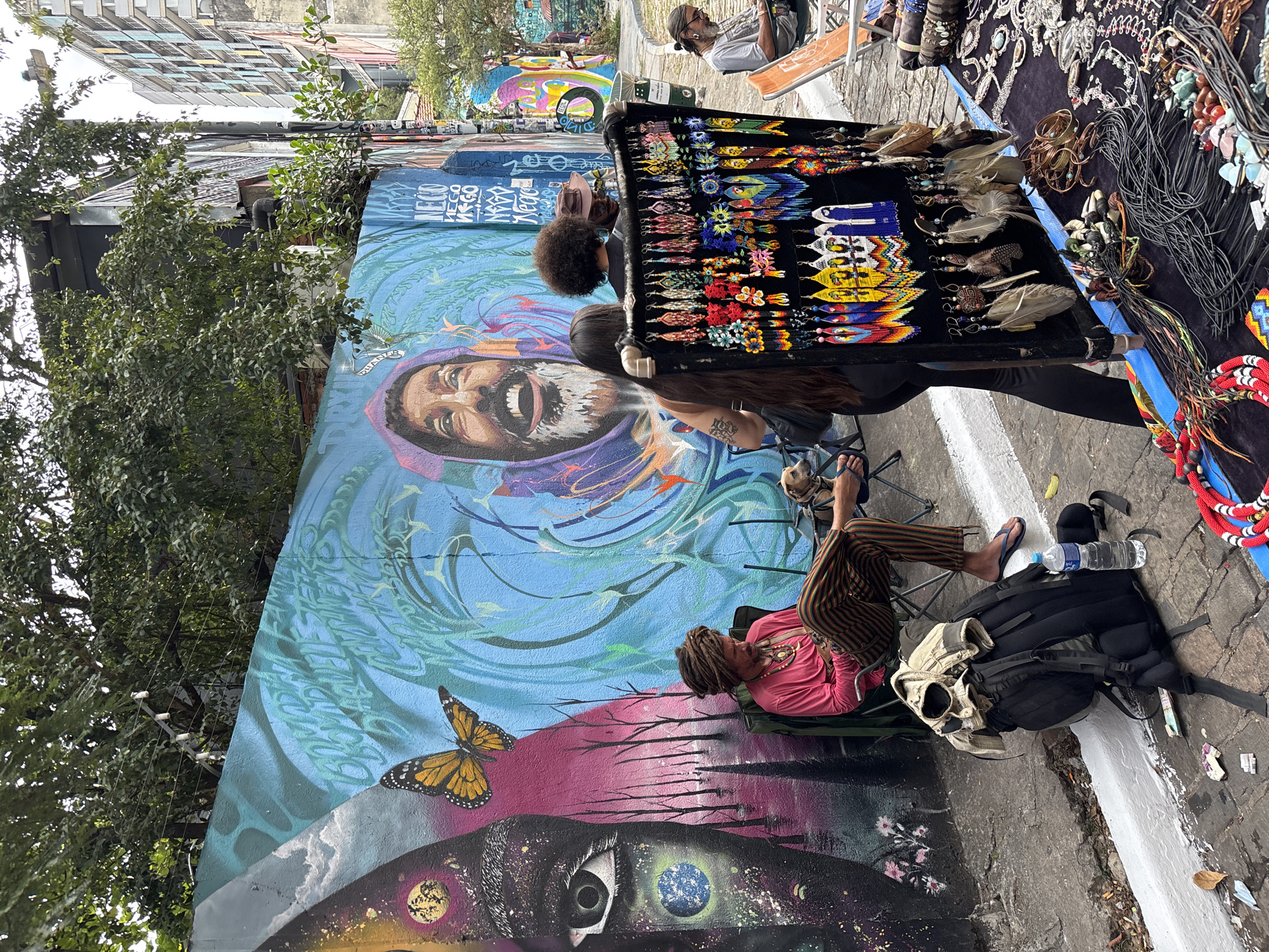 The image depicts a street scene in São Paulo, Brazil. A mural featuring a portrait of a bearded man, a butterfly, and a large eye covers a wall. Two individuals, one with dreadlocks wearing a pink shirt and striped pants, are seated in chairs next to the mural. A jewelry vendor stands behind a display table with various handcrafted earrings and necklaces. Other people are visible in the background. Backpacks and a water bottle rest on the ground near the seated individuals. The area appears to be an outdoor marketplace or an art district with pedestrian traffic.