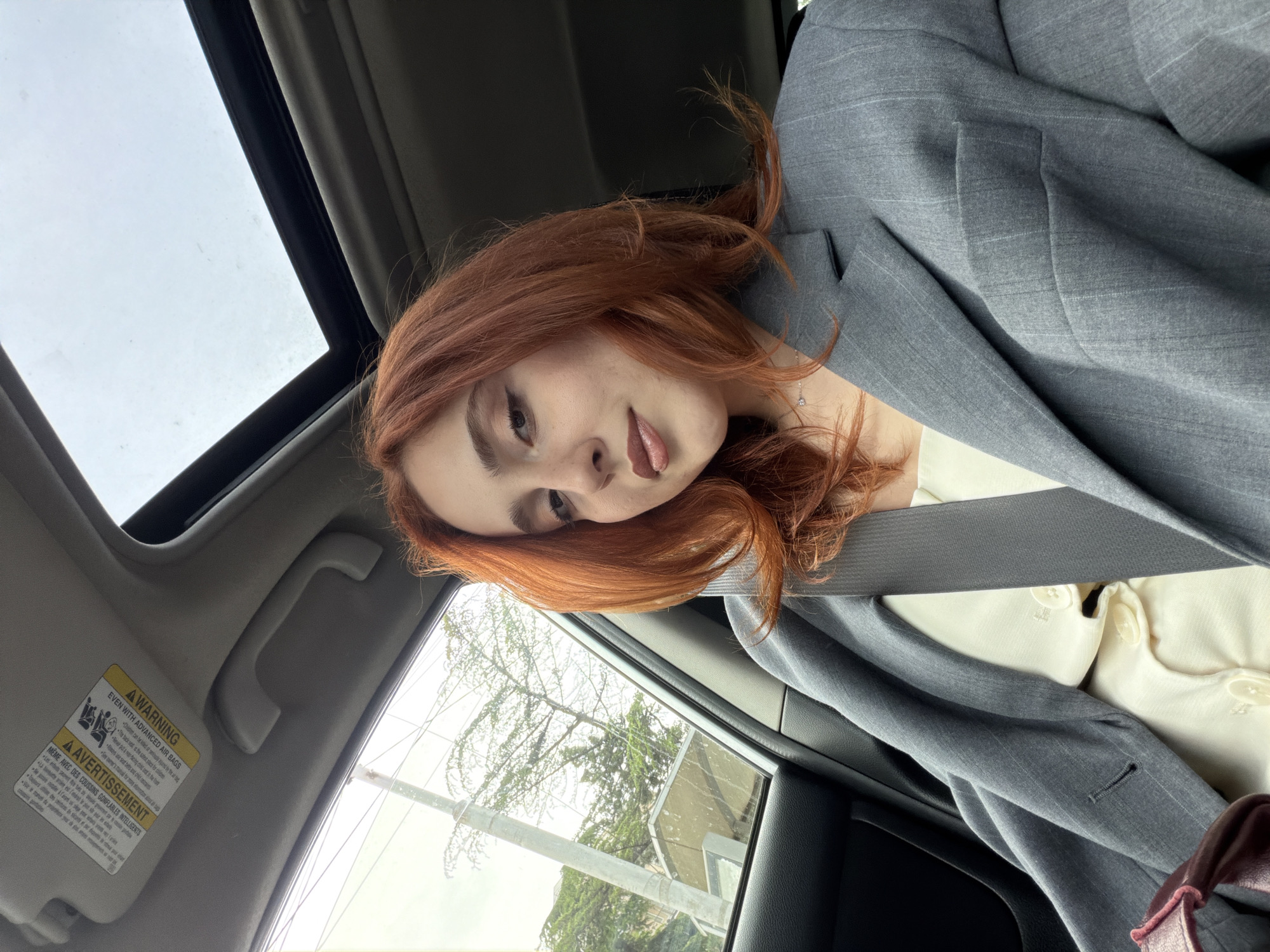 The image shows a red-haired woman sitting in a car, likely in Tbilisi, Georgia, given the presence of a warning sign in French (common in multilingual regions). She is wearing a grey blazer over a cream-colored top and is buckled into the seatbelt. The car has a sunroof, visible at the top of the frame. The view through the car window shows a tree and a building. The general scene suggests the woman is a passenger in a car traveling within the city.