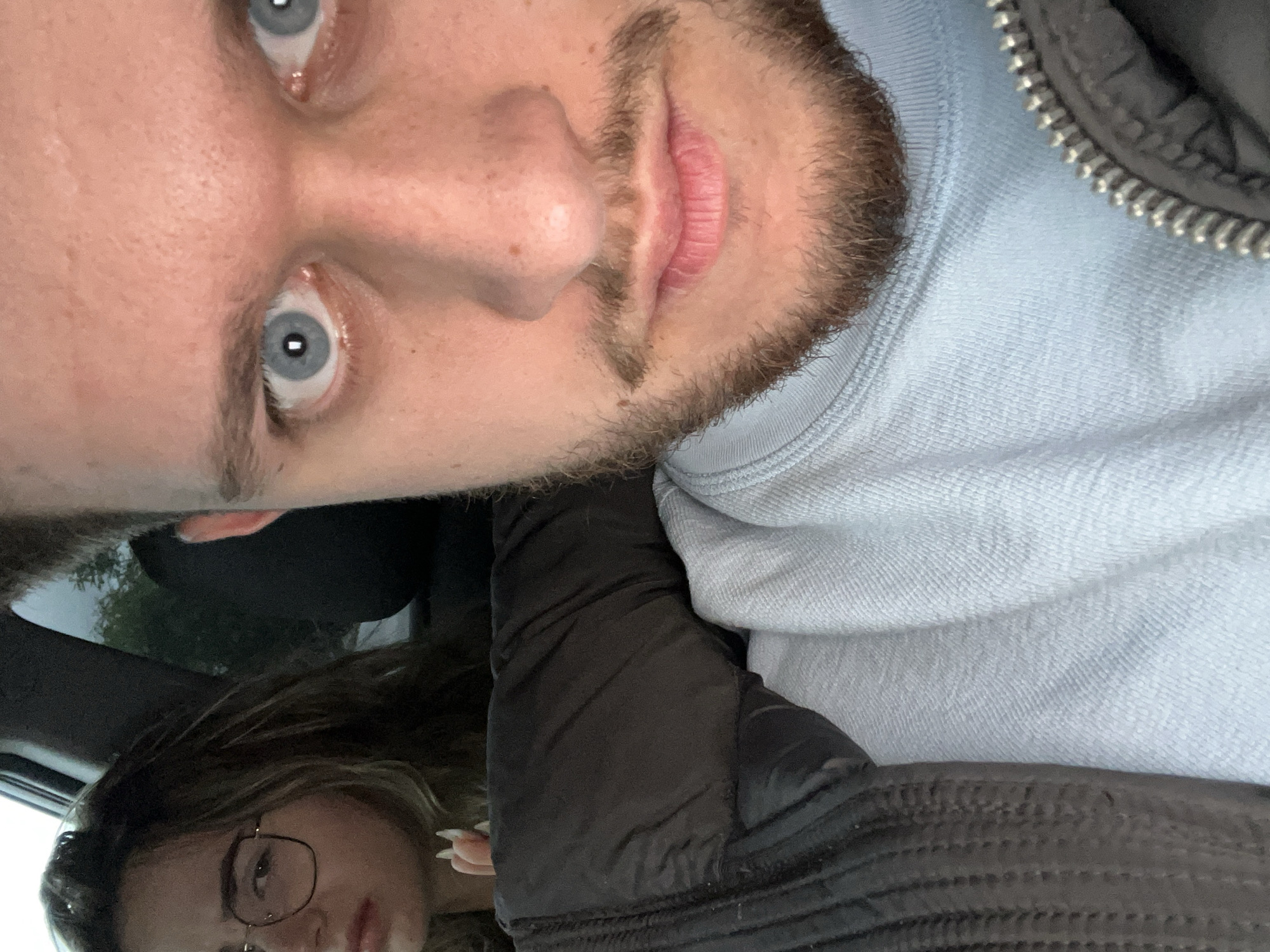 A selfie of a fair-skinned male with light blue eyes and a patchy brown beard. The man is wearing a light blue sweatshirt. Below him is a woman with brown hair and glasses, wearing a black puffy jacket. The location is inferred to be inside a car. Location is not discernible.