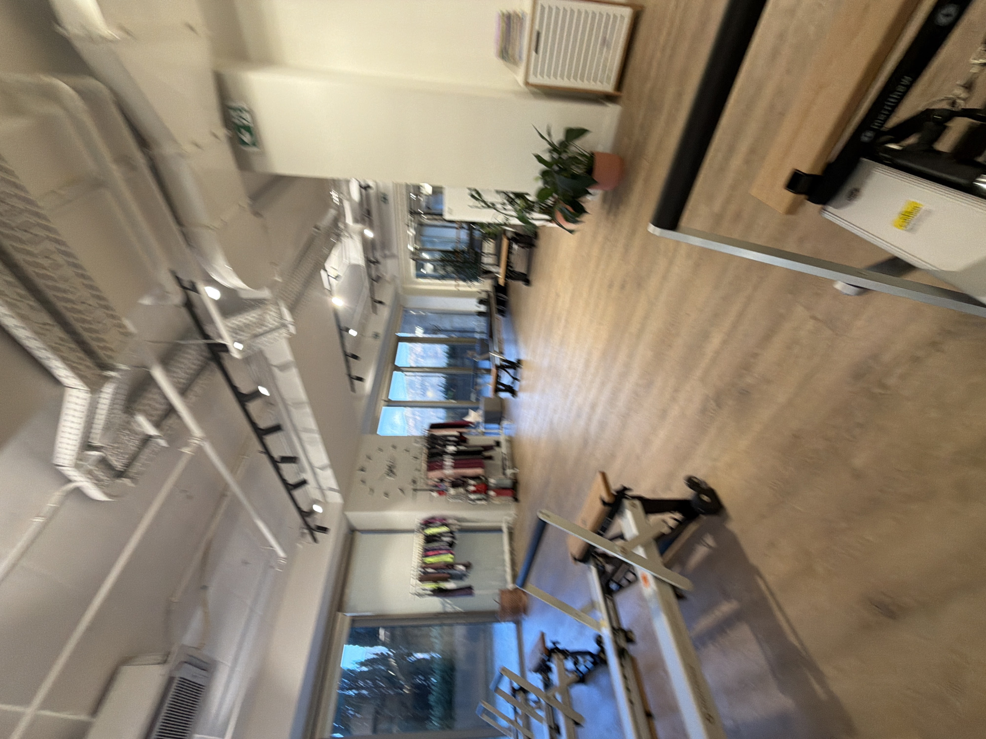 The image depicts the interior of a well-lit fitness studio, possibly a Pilates or yoga studio, situated in Boreti, Montenegro. The space features a polished wooden floor and large windows that allow natural light to stream in, offering a view of trees and the sky. Several pieces of exercise equipment, resembling reformers, are arranged in the foreground and mid-ground, each with a wooden surface and metal frame. In the background, racks of athletic clothing are displayed, suggesting retail or storage within the studio. The ceiling has exposed ductwork and lighting fixtures. A potted plant and a cabinet with a slatted door are also visible in the room. The overall impression is one of a modern, clean, and functional fitness environment.