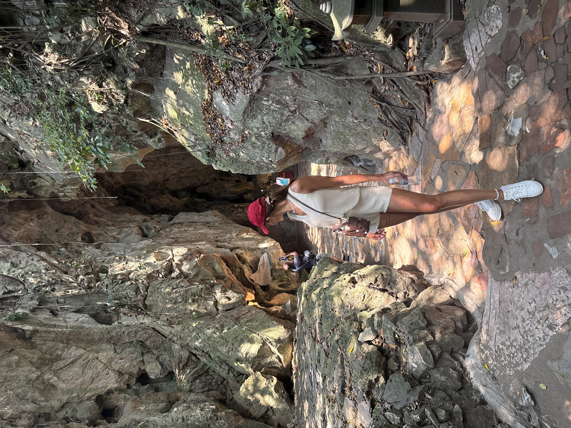 A woman wearing a red cap, white top, and shorts walks along a stone path in the Marble Mountains (Ngũ Hành Sơn) in Đà Nẵng, Vietnam. She is holding a water bottle and has a crossbody bag. Another person is visible further down the path among the rocks. The environment consists of natural rock formations and a stone-paved path.