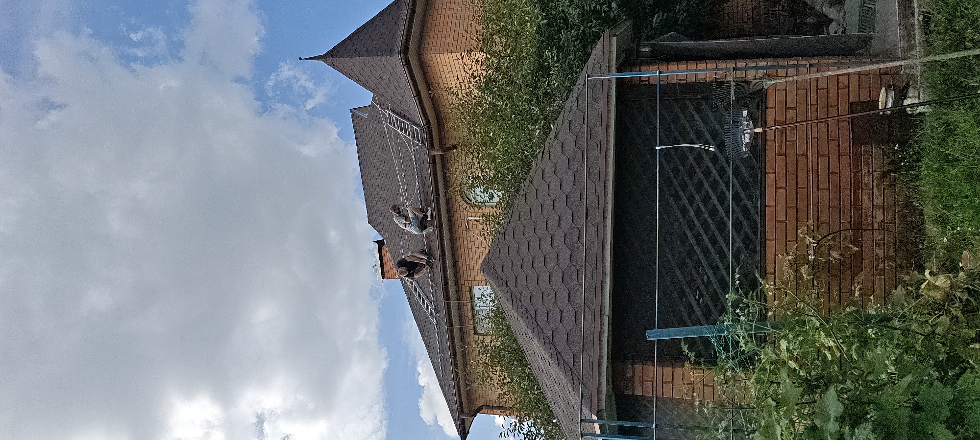 Two individuals are working on the roof of a brick house with a dark shingle roof. A ladder is propped against the roof, and ropes are stretched across it. One person, wearing a dark shirt and shorts, is kneeling and appears to be securing a rope. The other person, in a light-colored t-shirt and shorts, is also kneeling and looking down at the roof. The house has several windows and a prominent conical turret. In the foreground, a smaller brick structure with a dark shingle roof is partially visible, covered by dense green foliage. Two rakes are leaning against the brick wall of the smaller structure. The sky is partly cloudy with patches of blue. This scene is not located in Kyiv, Ukraine.