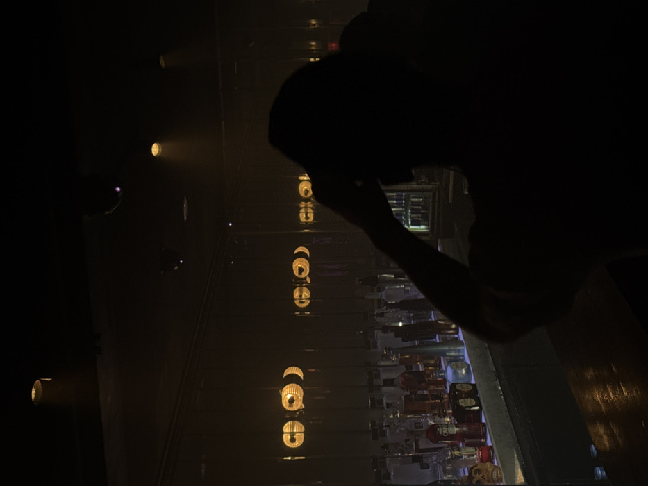 This image depicts an interior scene within a bar or nightclub located in Tel-Aviv, Israel. The environment is characterized by dim, warm, yellow-toned lighting. In the foreground, a heavily silhouetted individual, seen from behind and from the chest up, occupies the lower right portion of the frame. This individual faces away from the camera, towards a bar, with their right arm raised and hand positioned near the right side of their head, obscuring part of their profile.
Behind the individual, a commercial bar setup is visible. A dark countertop stretches horizontally across the lower-mid portion of the frame. Above the countertop, multiple shelves display numerous bottles of various alcoholic beverages, illuminated by an unseen light source that casts a soft, warm glow on their contents. Bottles containing clear, reddish, and amber liquids are discernible. A distinctive skull-shaped object, possibly a bottle or decorative piece, is positioned on the left side of the bar display. Metallic bar tools or shakers are also interspersed among the bottles.
The upper half of the background features a large, reflective surface, likely a mirror or glass panels, that reflects several overhead light fixtures. These reflections appear as bright, warm, circular or elliptical glows. The ceiling is dark, with visible recessed spotlights emitting concentrated beams of warm light downwards. On the far right, a vertical refrigerator or cooler with clear glass doors reveals additional beverage containers. No direct interactions between the silhouetted person and other individuals or objects are clearly observable, beyond the person's posture facing the bar.