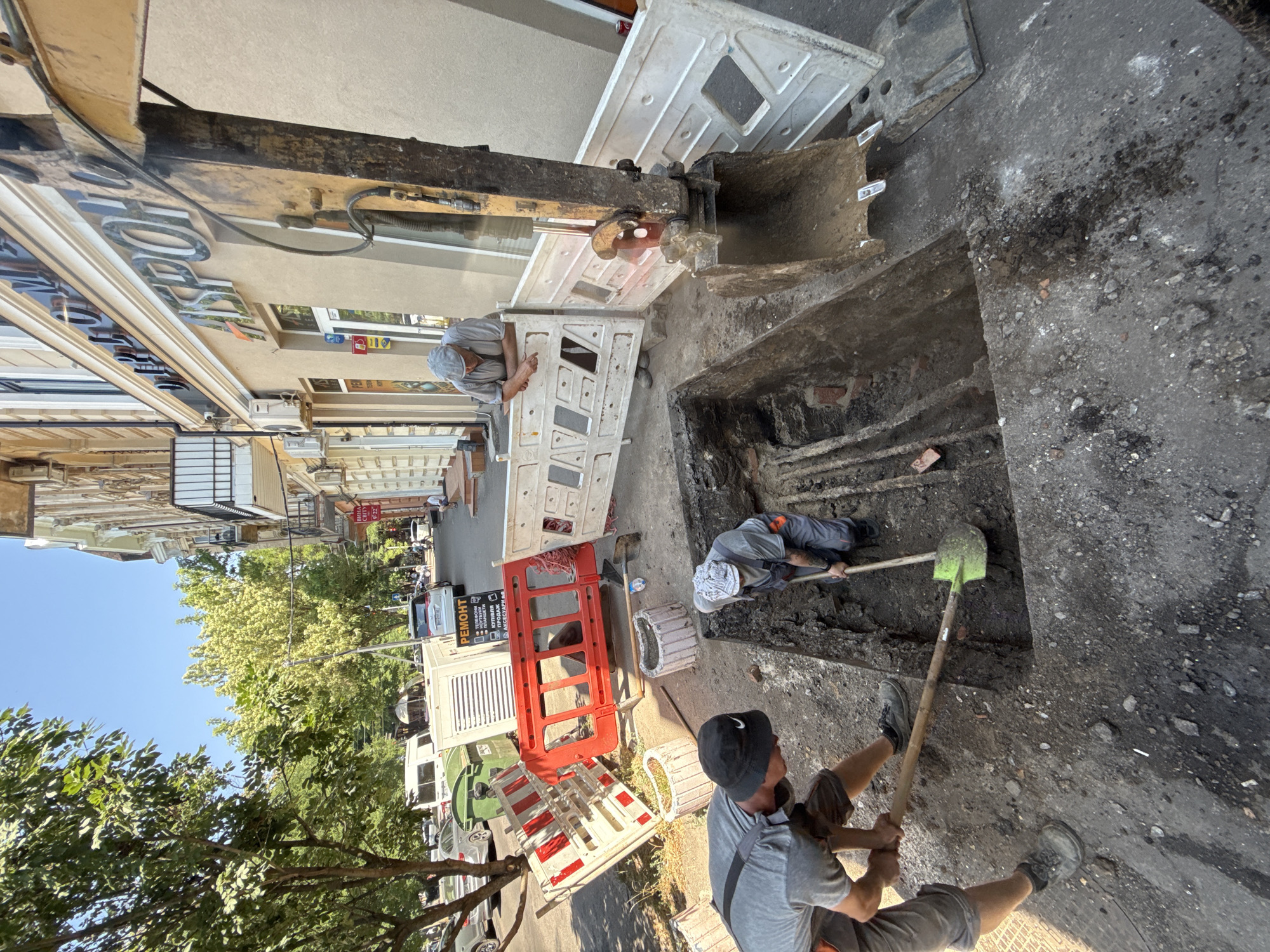 Three men are working on a construction site in a city. In the foreground, two men are digging a trench with shovels. One man is wearing a black bucket hat and a grey t-shirt with grey overalls, and is digging with a green-bladed shovel. The other man is wearing a grey cap and grey overalls, and is digging with a shovel. Behind them, a third man in a grey cap and t-shirt leans on a temporary barrier, observing the work. A yellow excavator is parked nearby, its bucket resting on the ground. The site is cordoned off with red and white barriers and white protective panels. Various construction equipment, including a portable generator, is visible in the background. The scene appears to be on a street in front of a building with an inscription above the entrance. The weather is sunny.