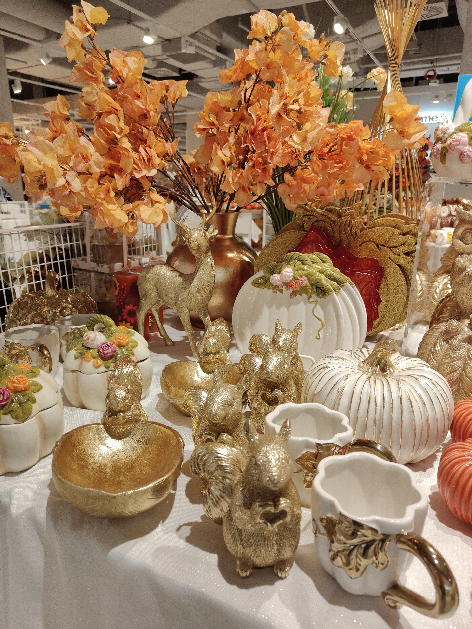 The image displays a collection of autumnal-themed decorative items arranged on a white tablecloth. Prominently featured are several ceramic pumpkins, some white with gold detailing and others ribbed orange. Scattered among them are golden squirrel figurines, a golden deer figurine, and gold decorative bowls. A large bouquet of vibrant orange artificial flowers in a gold vase dominates the upper left portion of the frame. In the background, shelving units and ceiling lights suggest an indoor retail environment. The scene is likely a display in a home decor store. No people are visible in the image. There is no information in the image to suggest it is located in Gdynia, Poland.