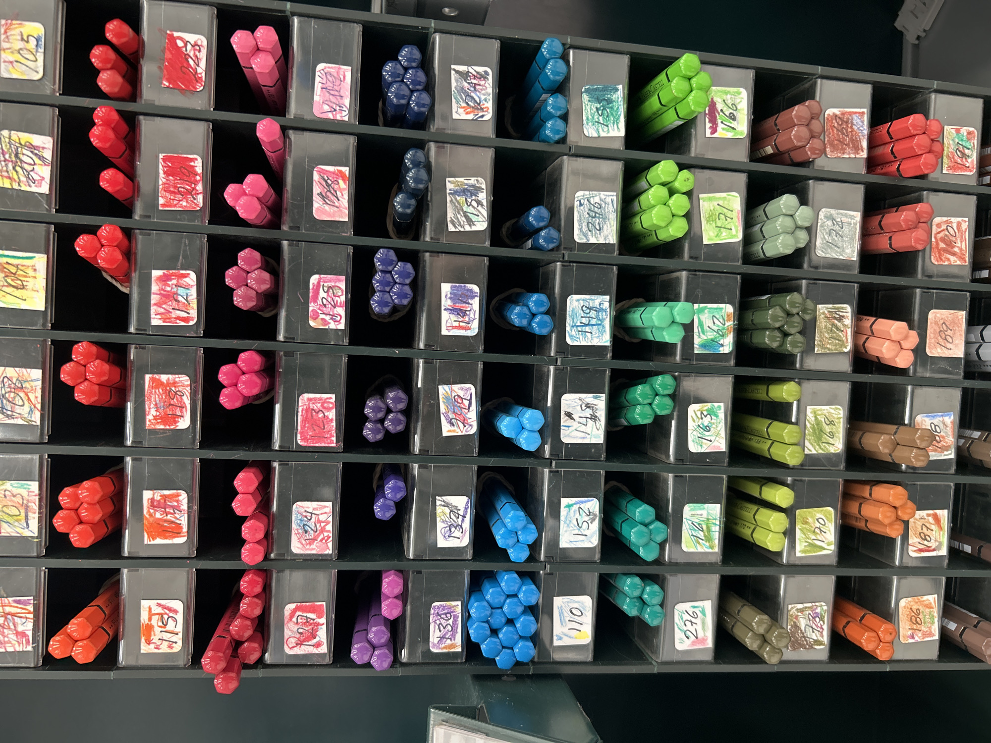 The image displays a collection of colored pencils or crayons organized within a storage unit composed of many small, individual compartments. The storage unit appears to be constructed of dark gray plastic.
Each compartment holds several pencils of a similar color, ranging from reds and pinks to purples, blues, greens, browns, and oranges. Each compartment also features a small, white label affixed to the front, which displays a number. Some labels are also adorned with scribbles, doodles, or other markings.
The pencils within each compartment are bundled together, suggesting they are pre-packaged or sold as a set. The arrangement of the compartments creates a colorful grid-like pattern across the frame.