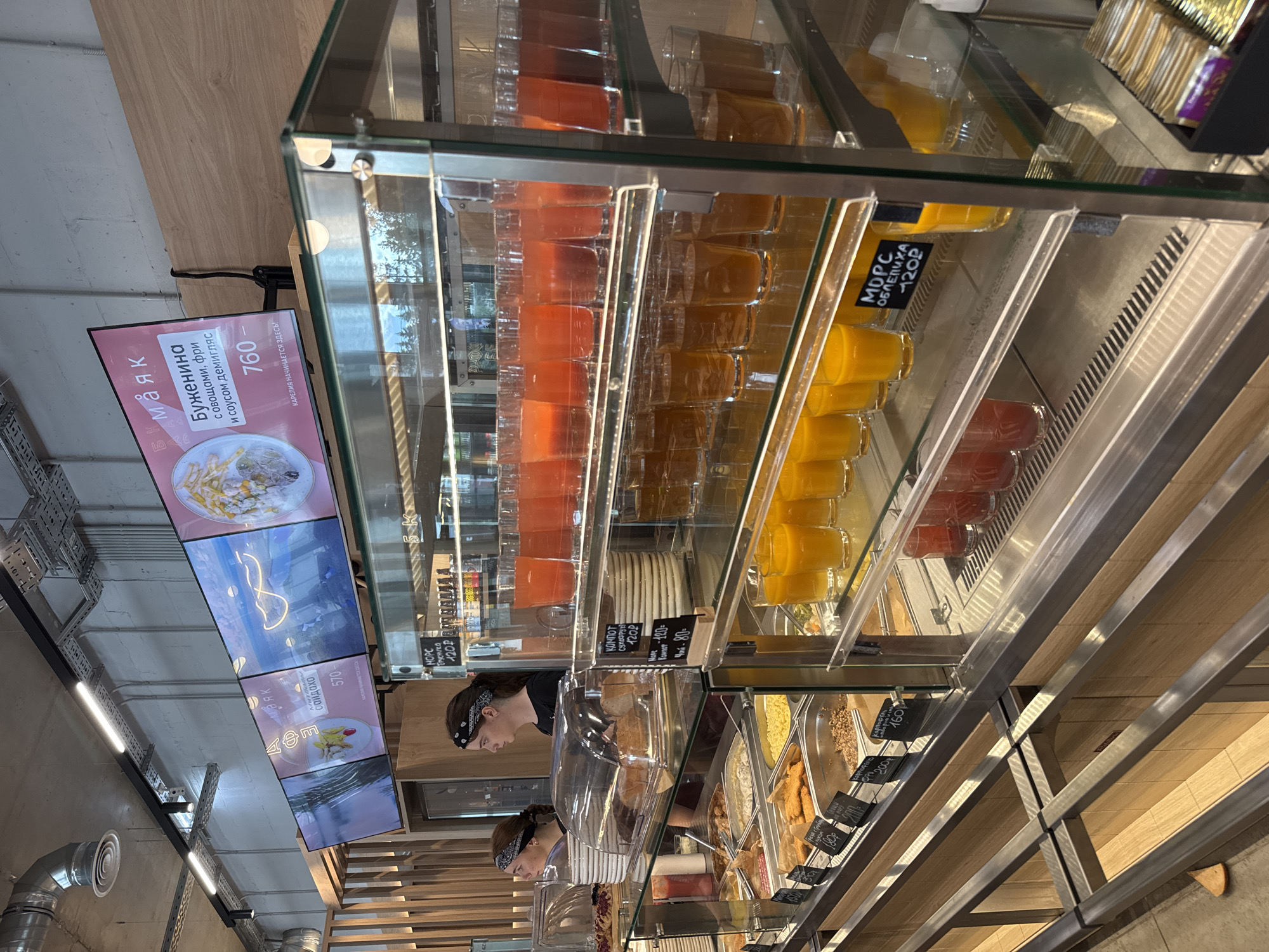 The image depicts the interior of a self-service cafeteria, likely in Yakkima, Russia, given the Cyrillic text on the menu boards and price tags. In the foreground, a glass display case filled with various food items, such as casseroles, fried chicken, and what appears to be grains, is visible. Several glasses of orange and red beverages are also displayed in the case, with price tags indicating "МОРС" (mors) and "КОМПОТ" (kompot). Two female employees, wearing bandanas, stand behind the counter, observing the scene. Above the counter, digital menu boards display food items and prices in Russian. The environment appears clean and well-lit, with industrial-style lighting fixtures and ventilation ducts visible on the ceiling. The overall impression is that of a modern, efficient food service establishment.