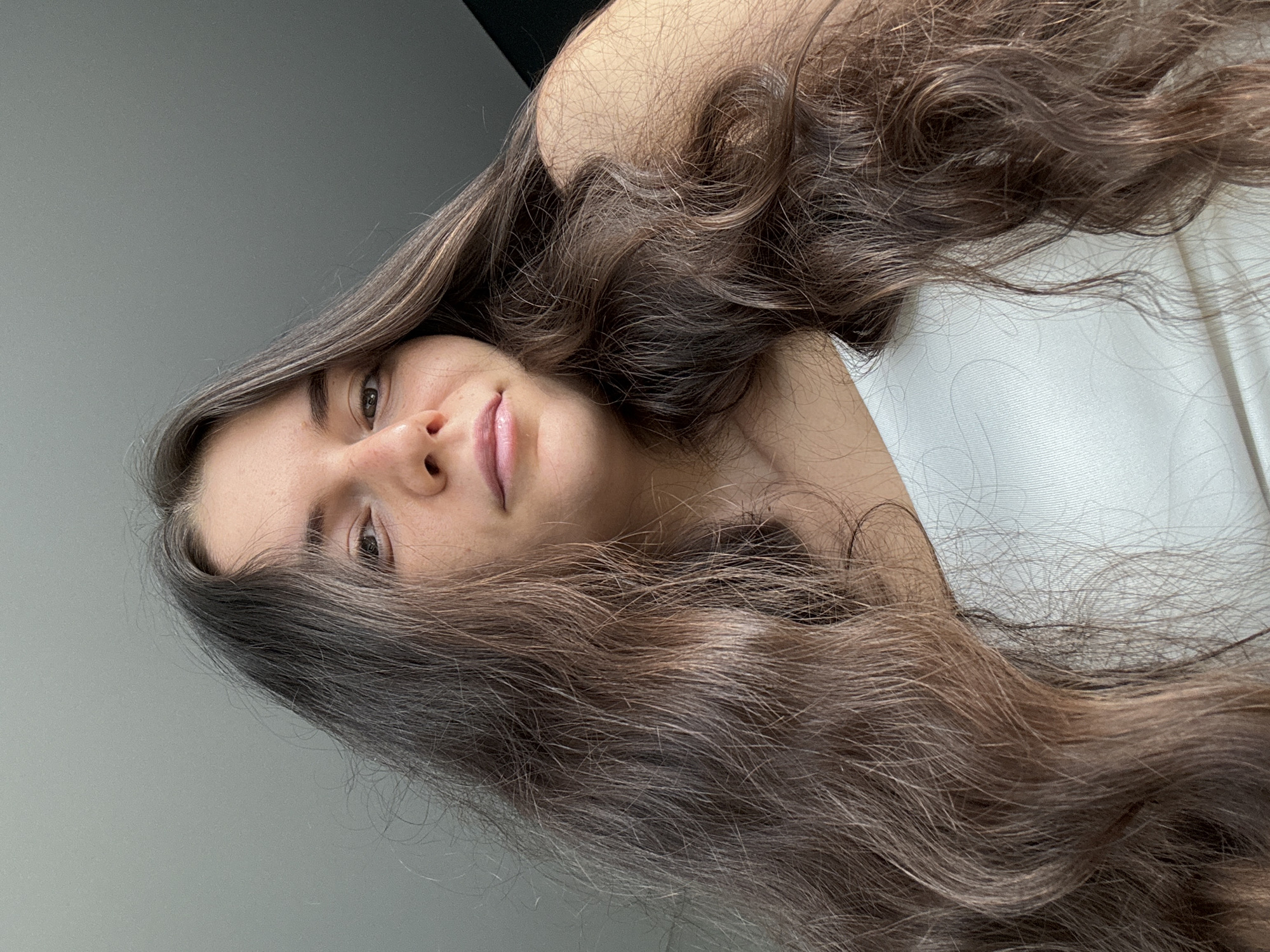A medium close-up shows a woman indoors, lying down with her head tilted at an angle. Her face is visible, with her hair flowing around her. She appears to be wearing a white top. The background is a light gray wall with a darker area in the upper left corner. No indicators point to Da Lat, Vietnam or any city specifically.