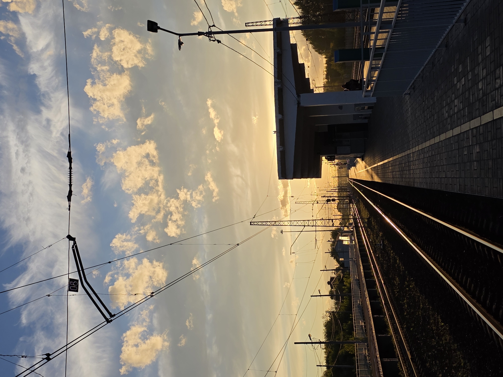 The image depicts a train station in Rechytsy, Russia, during sunset. The sky is filled with clouds that are illuminated by the setting sun, creating a warm, golden glow. Train tracks run through the foreground, leading towards the horizon. A train platform is visible to the right, with a covered waiting area and a streetlamp. In the distance, there are buildings and trees. The overall scene is serene and peaceful, conveying the beauty of nature and urban infrastructure.