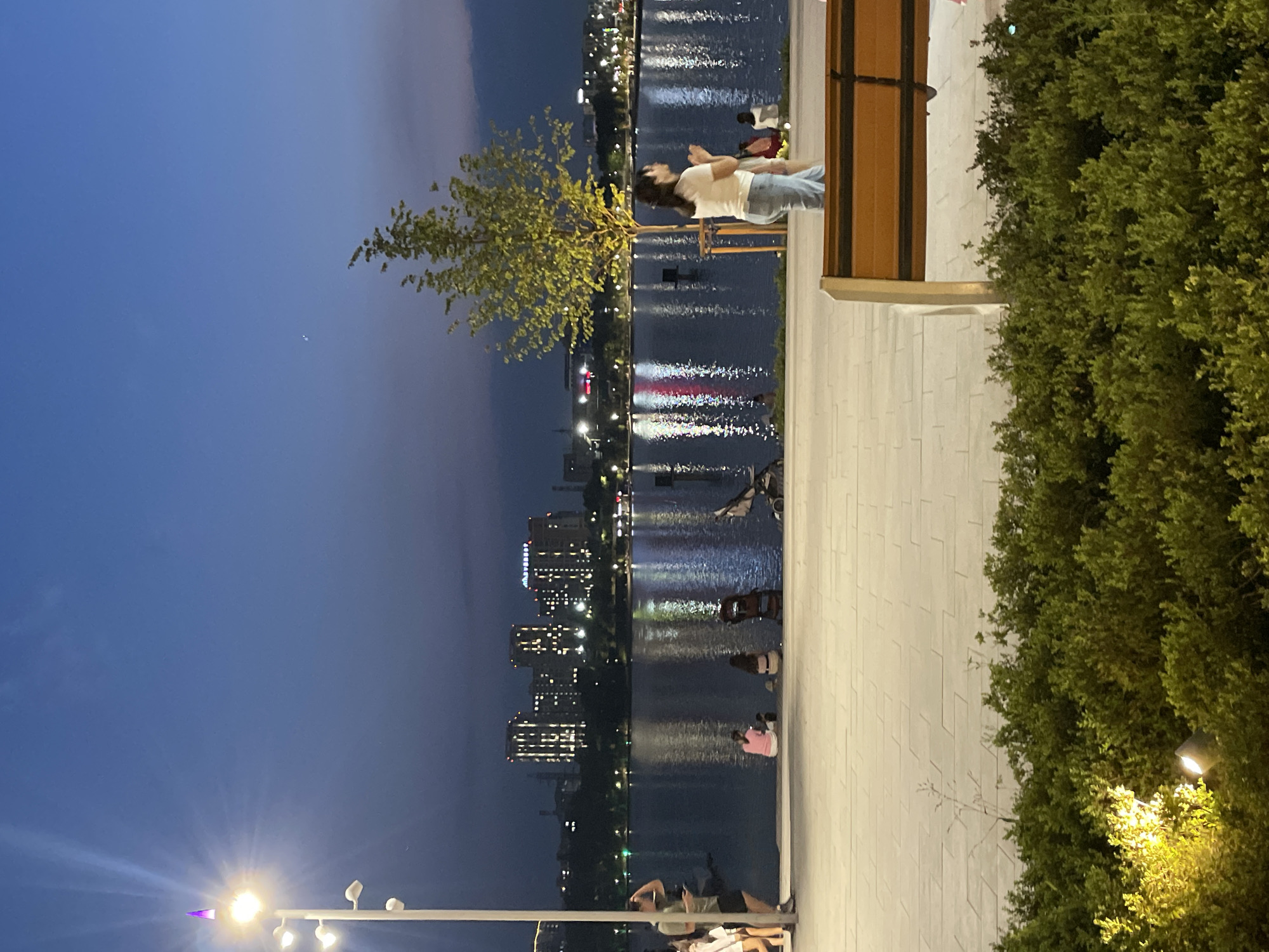This image depicts a dusk scene at the First President's Park in Almaty, Kazakhstan, along the banks of the Great Almaty Canal. The sky is a deep twilight blue, with a single bright star or planet visible. A modern lamppost with multiple lights, one topped with a purple beacon, stands on the left side of the frame. In the middle ground, the calm water of the canal reflects the lights of a distant cityscape, dominated by illuminated high-rise buildings. Several people are scattered along the paved walkway bordering the canal. On the left, a group of individuals are standing, and further along, several people are seated on the ground. A stroller is parked near the water's edge. In the right half of the frame, a woman in a white top and blue jeans stands facing away from the camera, appearing to be engaged in conversation or observation. A few other figures are visible further back near the water. A young tree with bright green foliage stands behind her. In the foreground, a section of a modern park bench with a wooden seat and metal frame is visible, set against a backdrop of dense green bushes, one of which is illuminated by a bright spotlight. The paving stones of the walkway are laid in a geometric pattern.