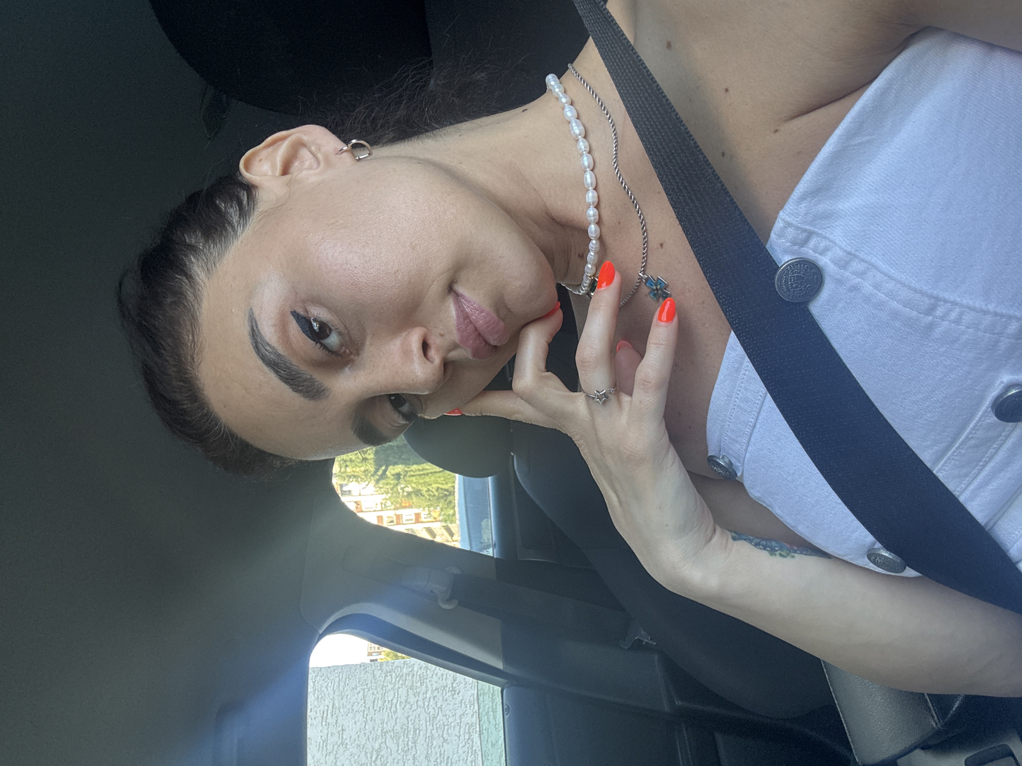 A woman with dark hair pulled back, wearing a white off-the-shoulder top, is seated in the passenger seat of a car. She has bright orange nail polish and is wearing two necklaces, one with pearls and another with a silver chain and a blue pendant. Her left hand rests on her chin, displaying a ring on her index finger. The interior of the car is visible, including a black seatbelt across her chest. Outside the car window, a building with white siding and some greenery can be seen. The image appears to have been taken during the daytime.