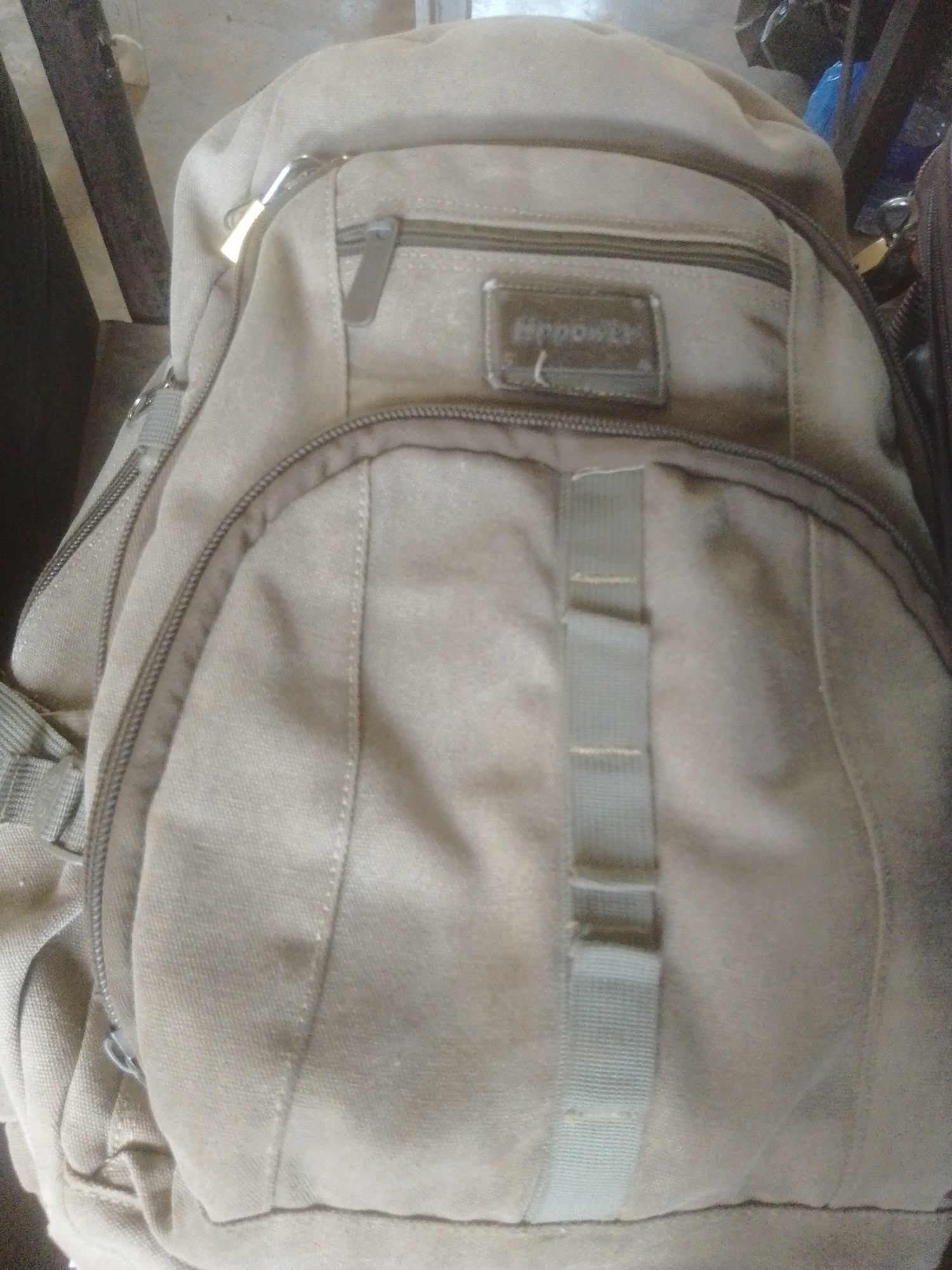 A tan canvas backpack is shown from a top-down, slightly angled perspective. The backpack has a main zippered compartment and a smaller zippered pocket on the front. A rectangular metal plate with the word "HPPONER" embossed on it is attached to the front pocket. Two green fabric straps with multiple loops are also visible on the front of the backpack. The backpack is resting on a dark, textured surface, possibly a floor or a bench. The background is out of focus but appears to be an indoor setting with some wooden elements and other objects. The image was taken in Danbazau, Nigeria.