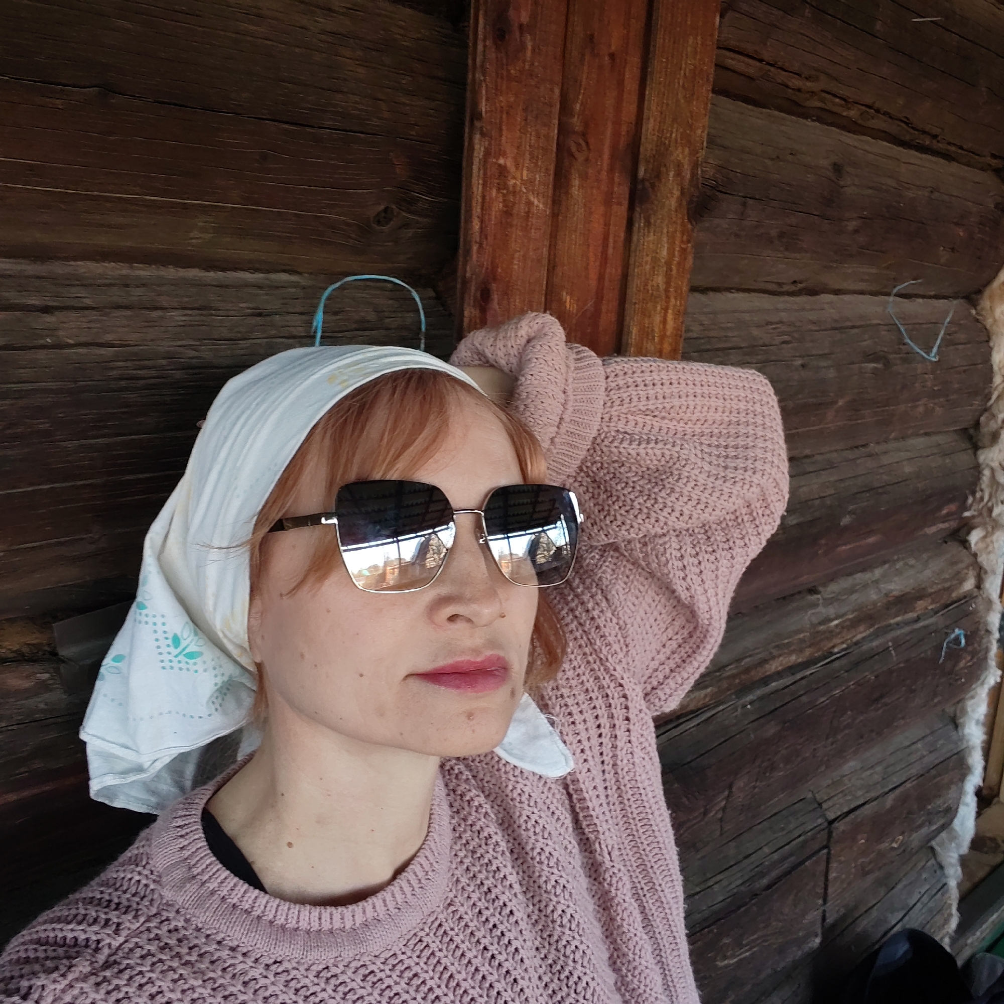 A woman with fair skin, wearing a headscarf and sunglasses, rests her head on a light pink sweater. She is positioned against a rustic wooden structure, likely a log cabin or building with exposed timber construction. The location is implied to be near Chaikovsky, Russia based on context, however, this cannot be definitively proven from the image.