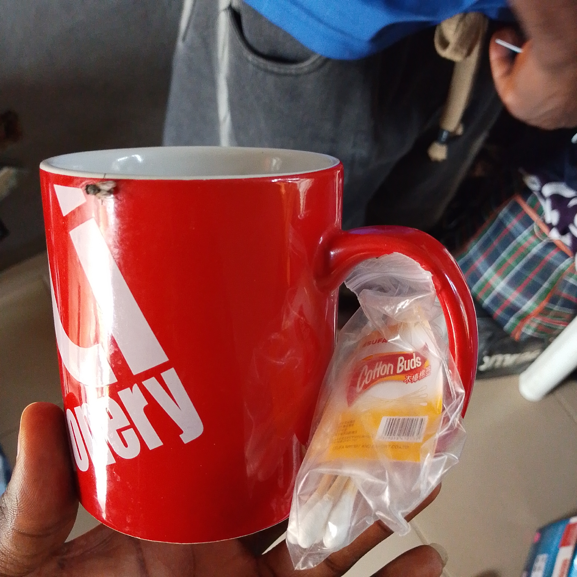 A close-up shot shows a red mug with white lettering on it, held by a person's hand. A few cotton buds in a clear plastic package are also visible, resting against the mug. A small insect is perched on the rim of the mug. In the background, a person wearing a blue shirt and jeans is partially visible. The setting appears to be indoors, possibly a casual environment. The lighting suggests it could be daytime. There is text visible on the cotton bud packaging, including "Cotton Buds" and some Chinese characters. The mug has white letters that appear to spell out "overy", with the beginning of the word obscured.