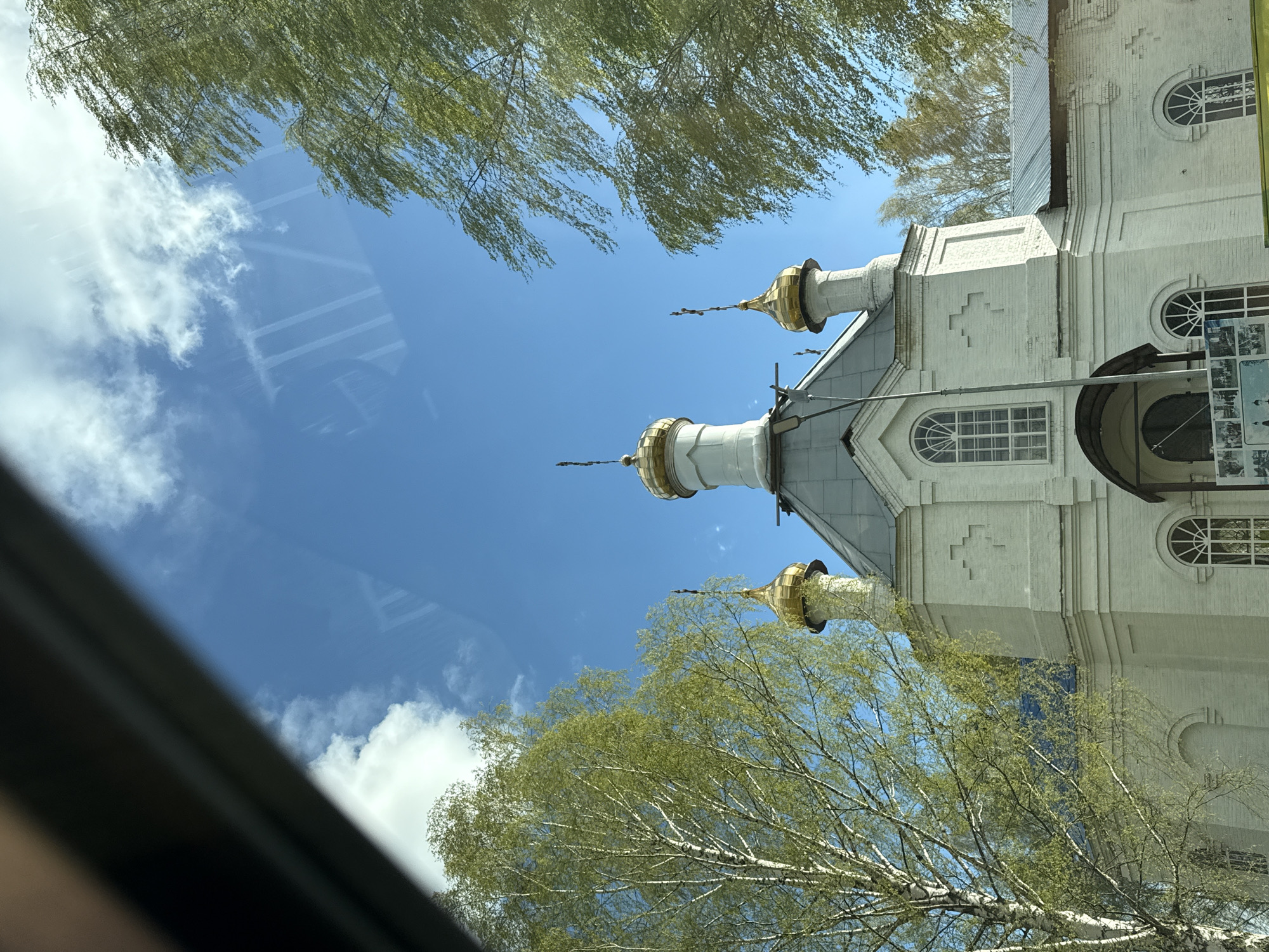 The image depicts a view of the Church of the Intercession of the Holy Virgin in Полозово, Russia. The perspective is from a low angle, looking upward through a window or possibly a windshield, as suggested by the partial frame visible on the left. The frame offers reflections of clouds and a bright blue sky. Above the frame, branches of birch trees with new green foliage are spread out. The church is a light-colored, likely white, brick building with three visible gold-domed towers. The architectural style includes arched windows and geometric decorations.