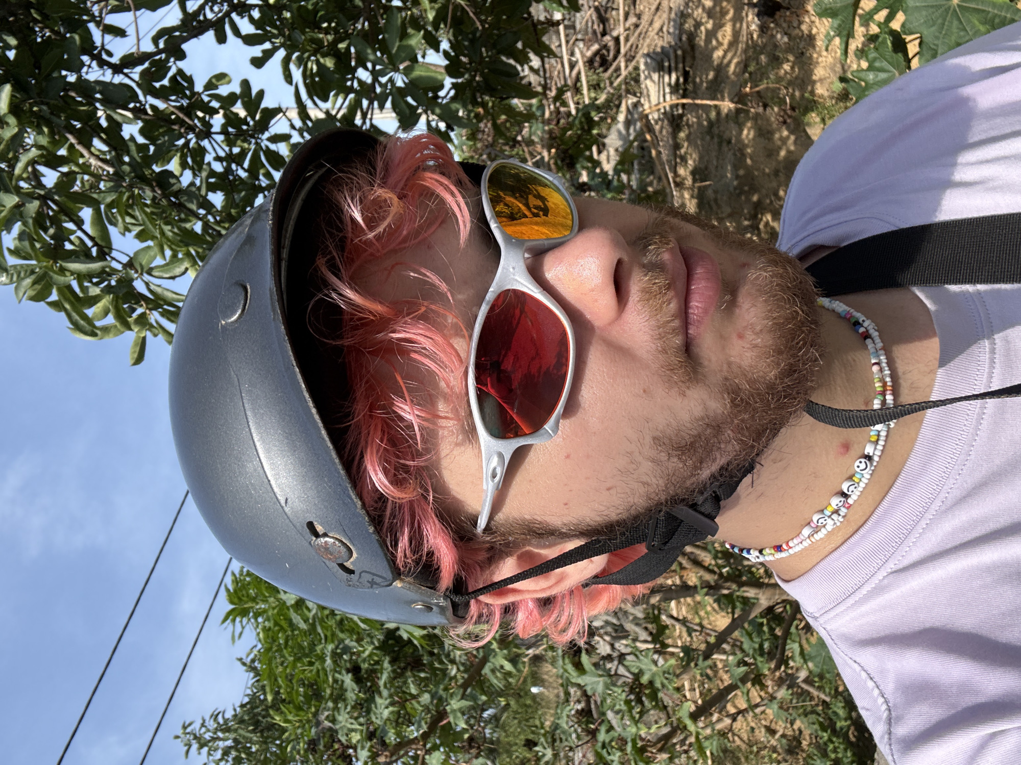 A person wearing a grey helmet, mirrored sunglasses, and a white t-shirt with beaded necklaces is the primary subject of the image. The person has pink hair and a beard. The background consists of foliage, trees, and a glimpse of a blue sky. Although there are other trees, a clear connection to Đà Nẵng, Vietnam is not evident in the image.
