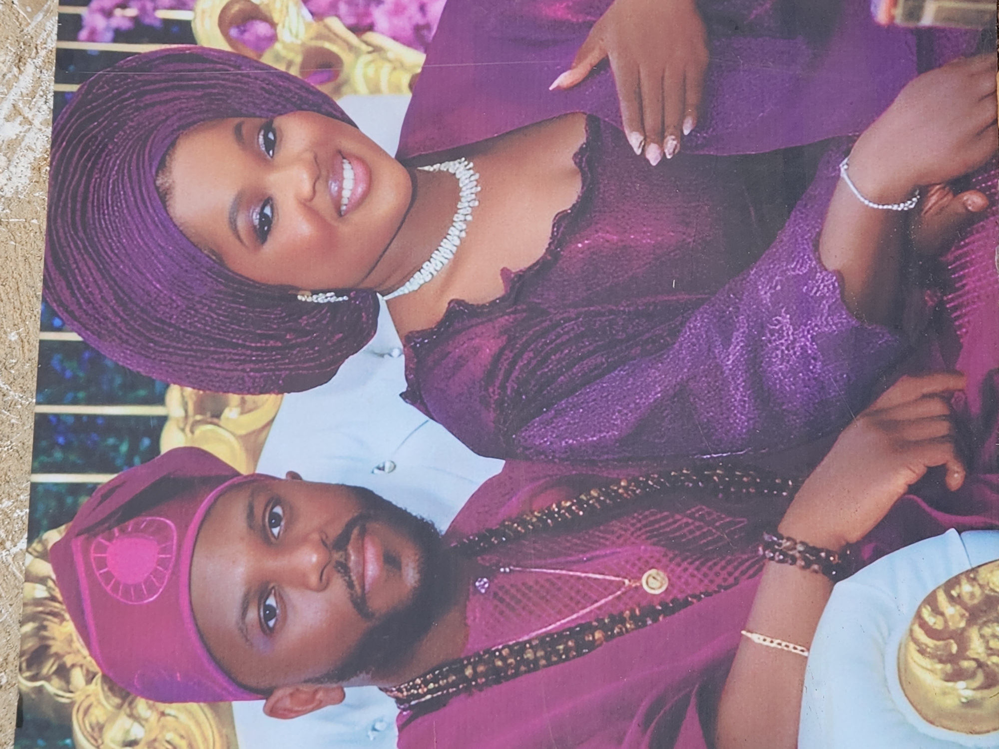 This image, set in Jos, Nigeria, features a smiling Nigerian couple formally dressed in traditional attire. The woman, positioned above the man and looking towards the viewer, wears a vibrant purple gele (headwrap) and a matching, shimmering traditional top. She accessorizes with a white beaded necklace, small white earrings, and a delicate silver-toned bracelet. Her nails are neatly manicured with a pink and white design.
The man, also facing the viewer, wears a deep purple fila (cap) with a distinct circular embroidered pattern and a coordinating traditional top. He is adorned with a multi-strand dark beaded necklace featuring a small circular gold pendant, a gold bracelet, and a dark beaded bracelet.
Both individuals are well-groomed, with the woman exhibiting a joyful, broad smile and the man maintaining a calm, composed expression. The background hints at an ornate indoor setting, featuring decorative gold elements and soft purple floral accents, suggesting a celebratory event like a wedding or engagement ceremony. The image appears to be a photograph of a printed picture. No visible text is present.