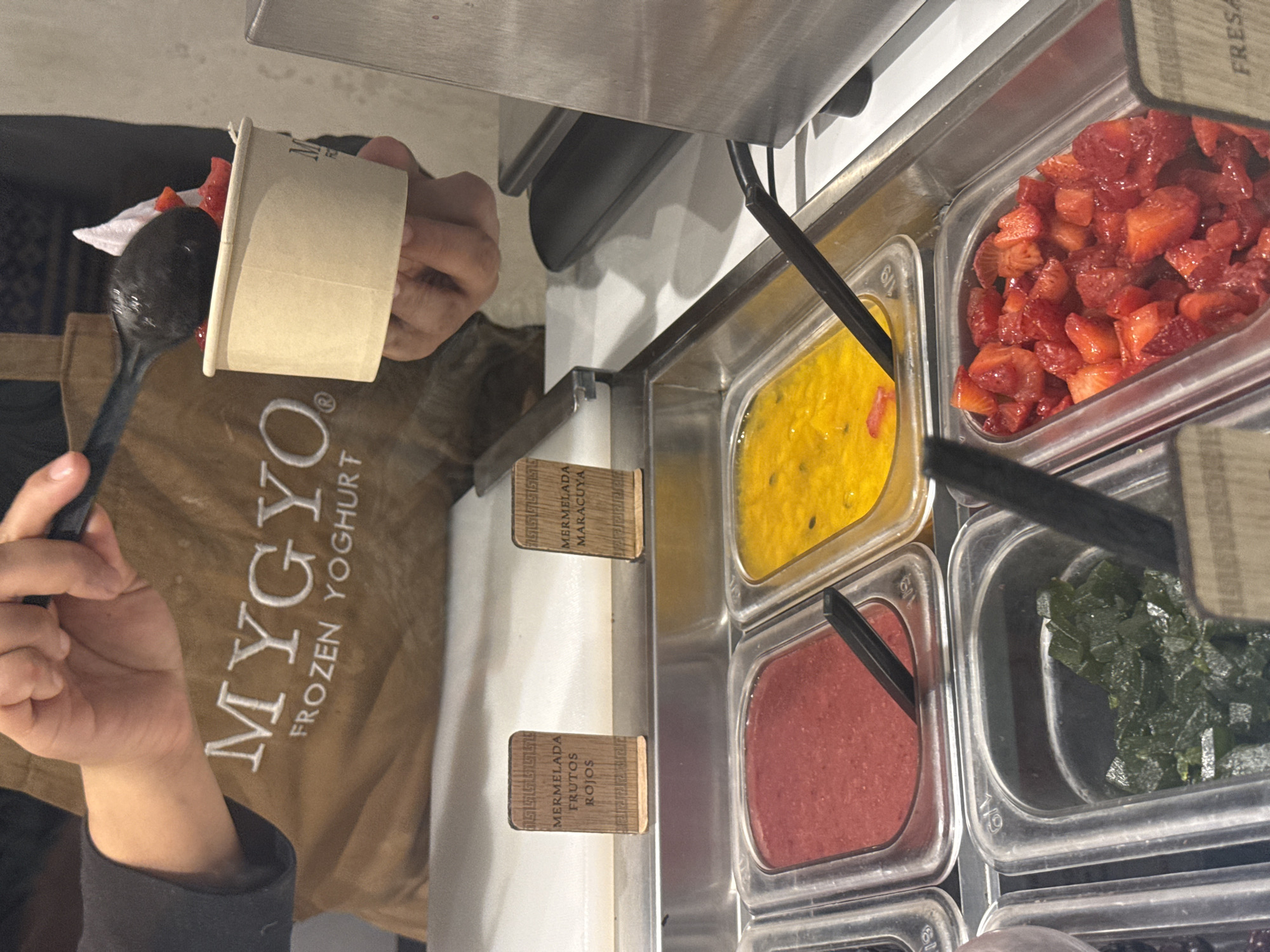 A person wearing a brown "MYGYO FROZEN YOGHURT" apron is holding a brown paper cup and a black spoon with diced strawberries. The topping station contains a variety of toppings, including diced strawberries, "mermelada maracuya" (passion fruit jam), "mermelada frutos rojos" (red berry jam), and diced green gelatin. The location is likely a Mygoyo Frozen Yogurt shop in Mexico City, Mexico.