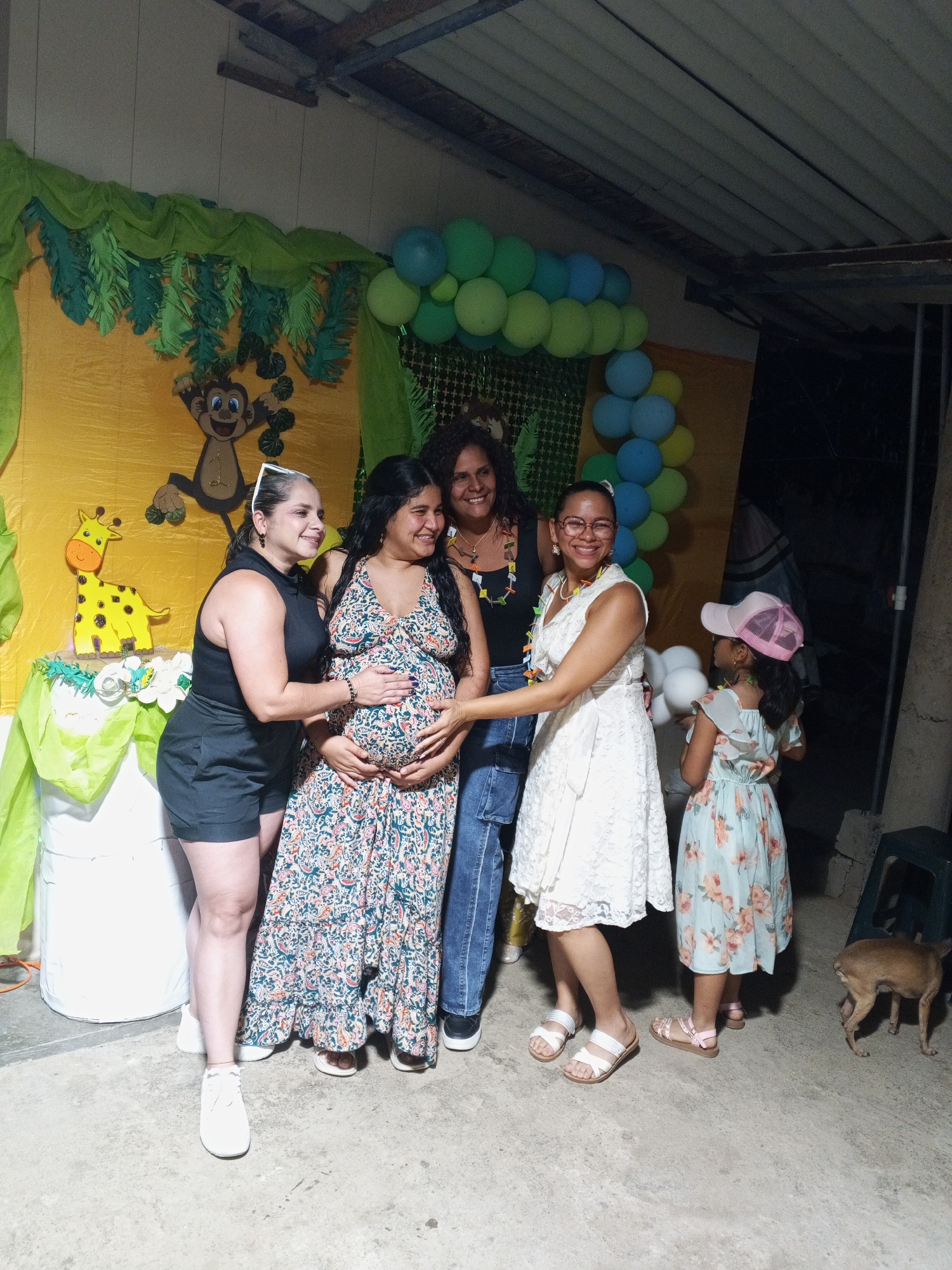 The image shows five people, including a pregnant woman, posing for a photo in front of a backdrop decorated with a jungle theme including animal cutouts and balloons. The pregnant woman is wearing a patterned dress. The woman on her left is wearing a black outfit and white sneakers. To the right of the pregnant woman, two women are smiling. A young girl in a floral dress and pink hat stands slightly behind them. A dog is visible to the right in the frame. The location appears to be an outdoor setting in Unknown, Unknown.