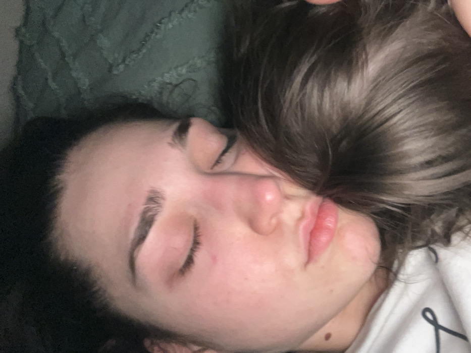 A woman is lying on her side, eyes closed. She has dark hair and fair skin with slightly flushed cheeks. Her eyebrows are full and dark. She has full lips and a mole on her neck. Her hair is partly obscuring her face. She is wearing a white top with black lettering. The background includes a textured green surface, possibly a blanket or upholstery.
