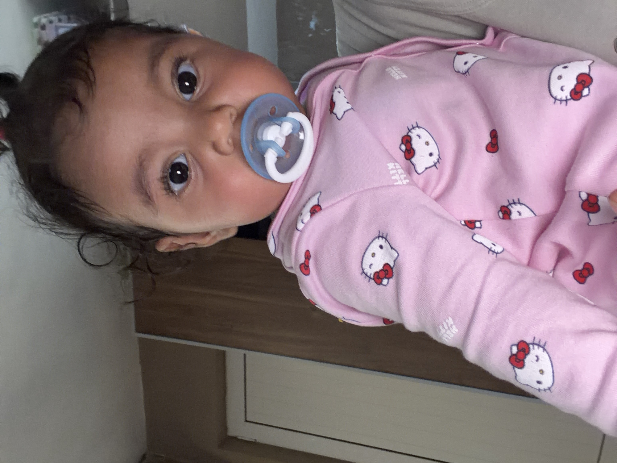 A low-angle, close-up, medium shot captures a baby wearing a pink Hello Kitty pajama set. The baby has dark hair pulled up and is looking directly at the camera with large, dark eyes. A blue pacifier is in the baby's mouth. The background consists of a light brown wall and a white door. The image appears to be taken indoors. The city of Razhevo Konare, Bulgaria is not depicted in this image.