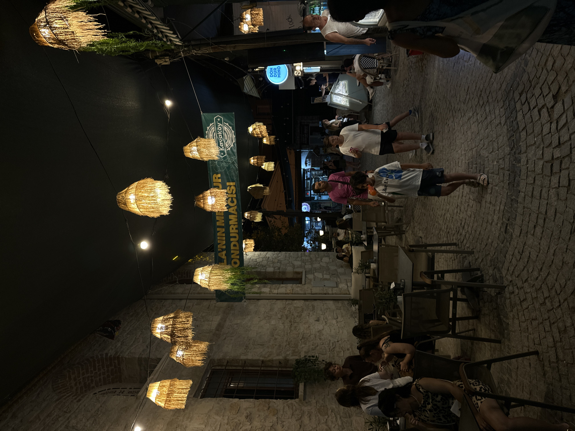 The image depicts a nighttime scene on a cobblestone street in Urla, Turkey, likely in a pedestrianized area with outdoor seating for businesses. Several people are present, some seated at tables and others walking. The street is illuminated by hanging pendant lights with fringed shades and a few other overhead lights. The architecture visible includes a stone building on the left with a large window. A banner with Turkish text, including "Kavaköy Dondurma" which translates to "Kavaköy Ice Cream," is hung across the street, suggesting the presence of an ice cream shop. In the background, more shops and businesses are visible, along with more people. The overall atmosphere is that of a lively evening gathering place.