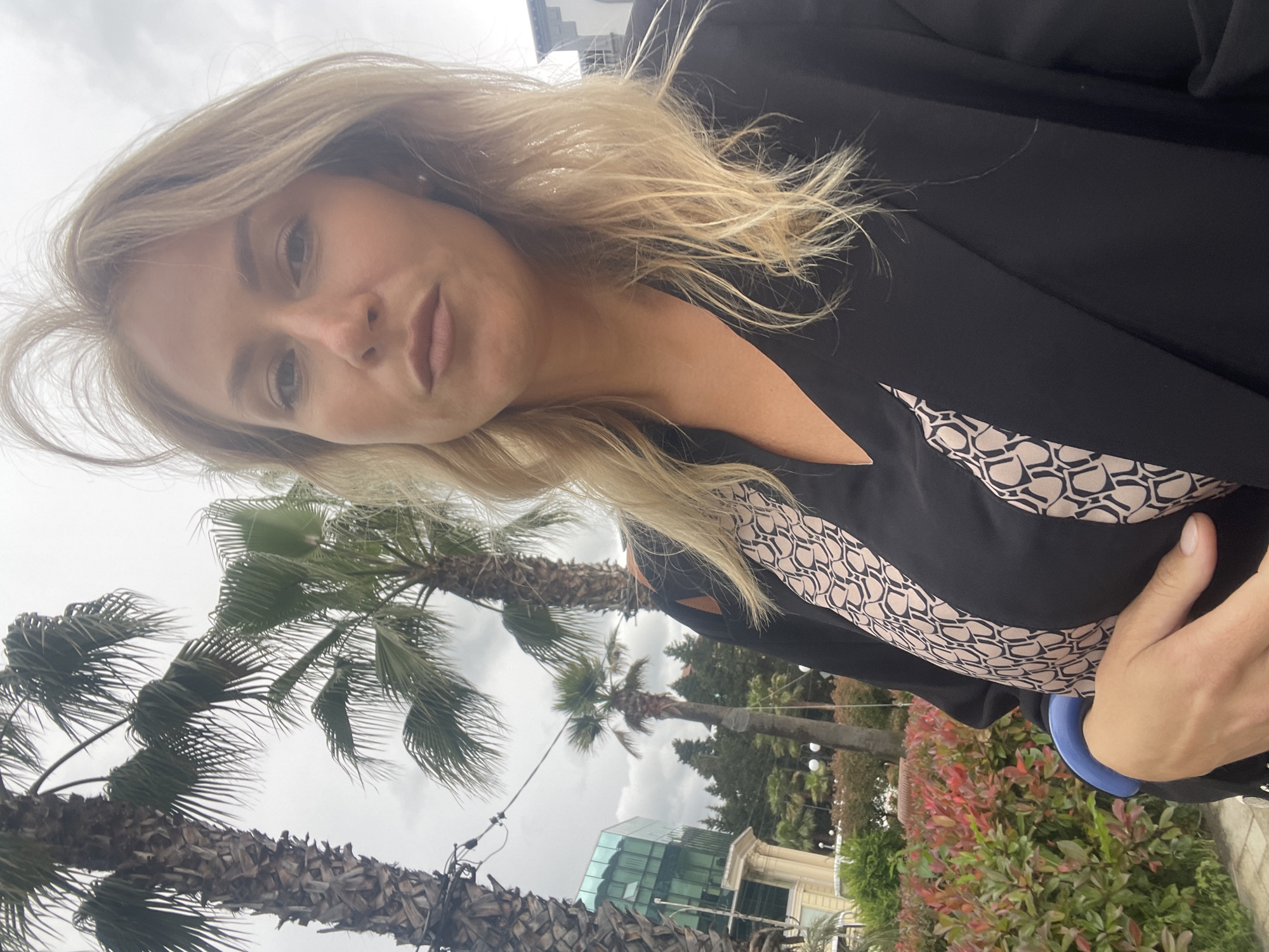 A selfie photograph shows a blonde woman in a black blazer with a patterned top, standing outdoors in Sochi, Russia. Palm trees are visible in the background, along with other greenery and a modern glass building. The sky is overcast. The woman is wearing a blue bracelet.