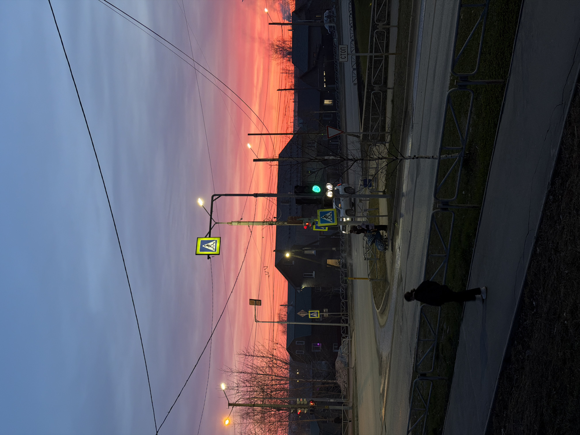 This image captures an urban street scene at twilight, likely sunset, characterized by a strikingly colorful sky. The horizon glows with intense reds and oranges, transitioning upwards through shades of pink and purple to a lighter blue-grey. Faint cloud formations are visible against this dramatic backdrop.
In the foreground and midground, a paved road with sidewalks runs through the frame. Several dark-silhouetted buildings, possibly houses or low-rise structures, stand against the vibrant sky. Streetlights are illuminated, casting warm glows, and traffic lights are visible, with one showing green and another red. Utility poles and a network of overhead wires crisscross the upper portion of the scene.
Prominently displayed are multiple yellow square signs with black pedestrian symbols, indicating pedestrian crossings. One white rectangular sign clearly displays the Cyrillic word "СТОП" (STOP). Metal fences line the sidewalks and separate them from grassy areas. Bare trees, possibly in late autumn or winter, are scattered throughout.
One person, dressed in dark clothing, is walking on the sidewalk in the lower right quadrant, moving away from the viewer. Another figure, possibly pushing a stroller or wheelchair, is faintly visible near a crosswalk further down the street. A few vehicles are present on the road. The overall setting appears to be a residential or mixed-use area within Berezniki, Russia, as suggested by the Cyrillic text.