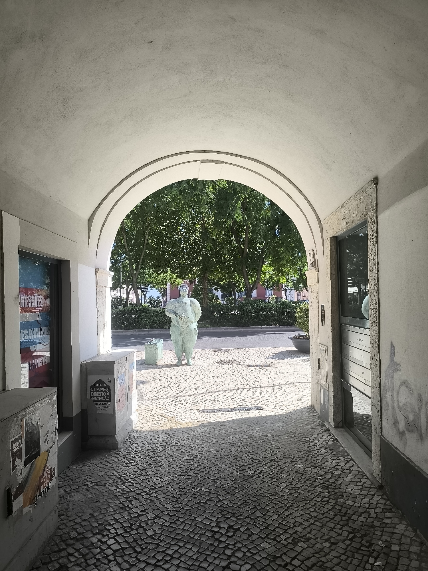 The image depicts an outdoor scene viewed from within a covered walkway or archway. The walkway has a white, arched ceiling and white walls. The floor of the walkway is paved with grey cobblestones. To the left, there is a glass-fronted storefront with posters visible inside and on the exterior wall. A grey metal bin with more posters is also on the left.
Exiting the archway, a street paved with cobblestones is visible. On the street, a bronze statue of a stout figure holding a camera stands. Next to the statue is a green trash receptacle. Lush green trees and vegetation line the street in the background. The lighting suggests it is daytime, with shadows cast by the archway and the statue. The statue is located in Praça de Bocage in Setúbal, Portugal.