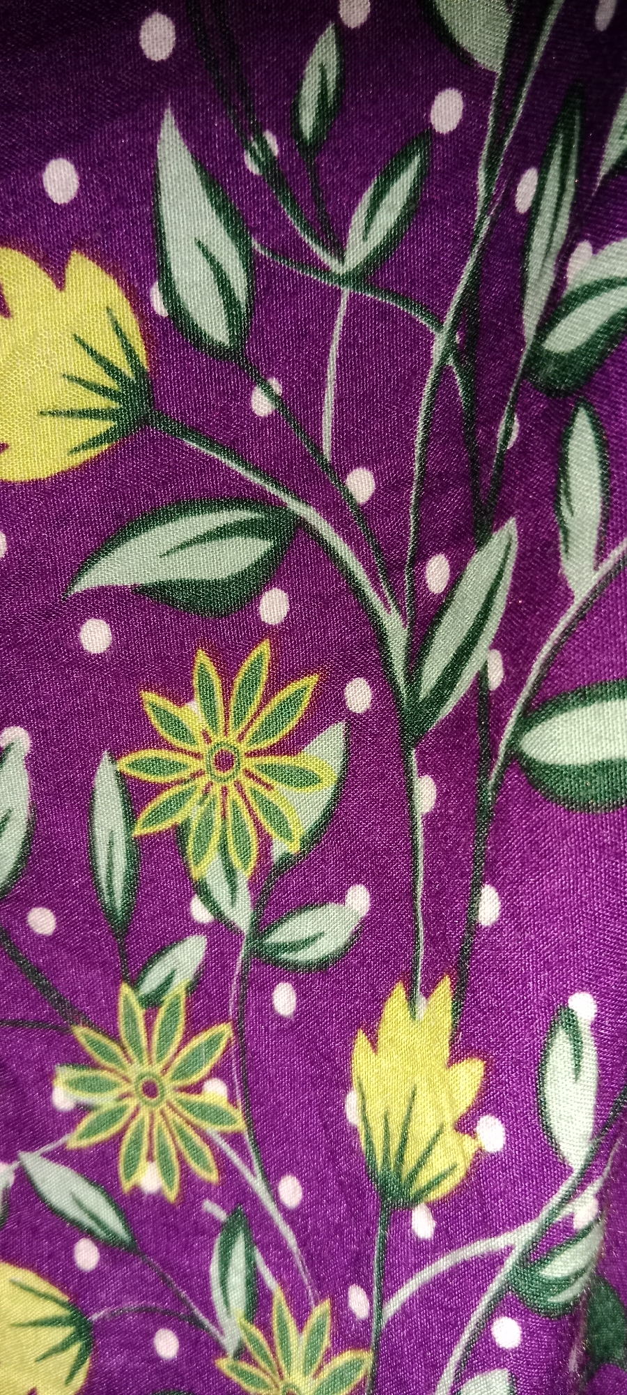 The image displays a close-up view of a purple fabric with a floral pattern. The pattern consists of stylized yellow flowers with green outlines, and green leaves scattered on the purple background. Small white dots are interspersed throughout the fabric, adding to the decorative design. The fabric appears to be a textile material, possibly from clothing or upholstery. The image does not contain any information or context related to the city of Subang, Indonesia, or any outdoor environment.