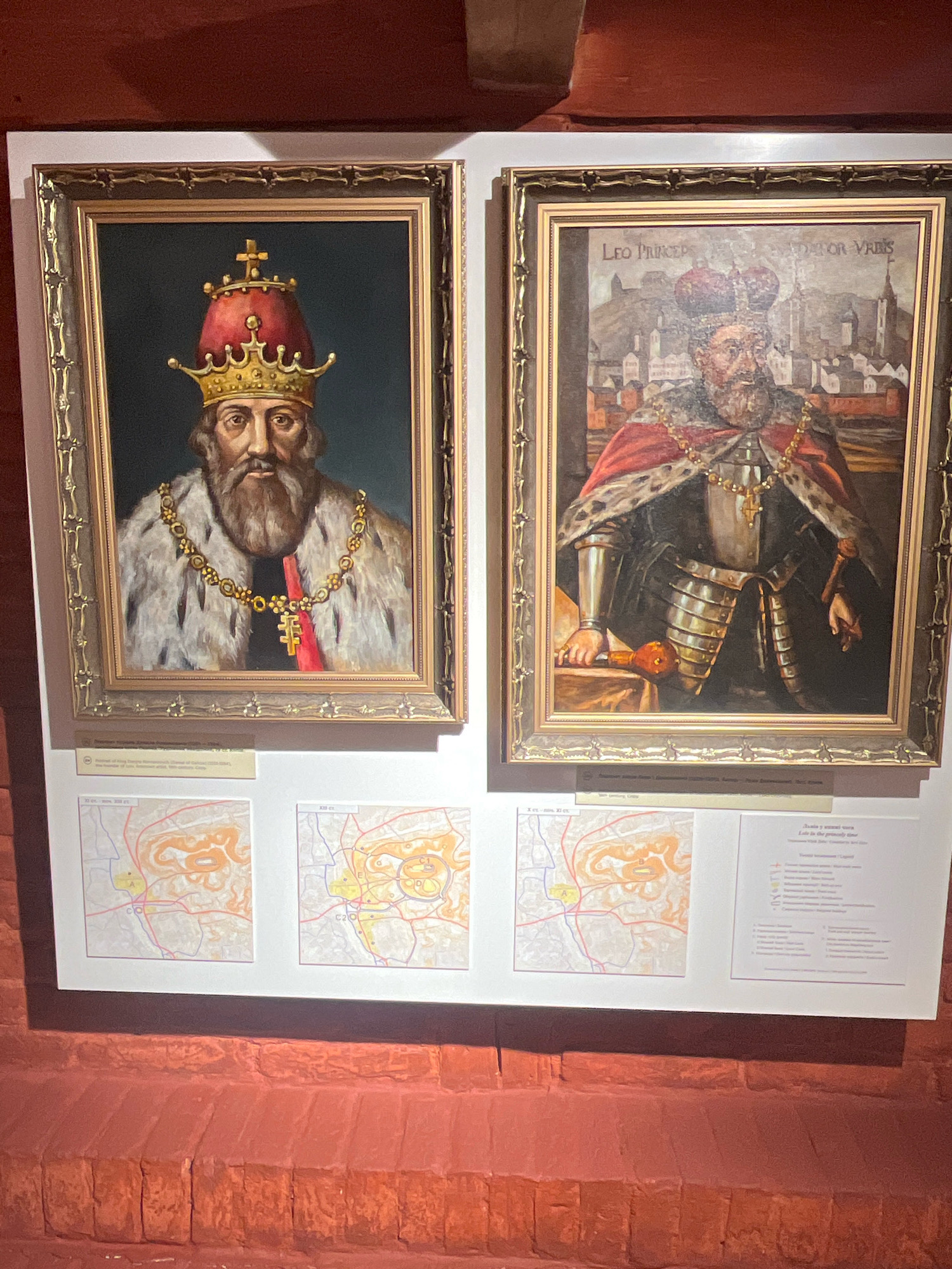 The image displays two framed oil paintings of historical figures, likely rulers, mounted on a white wall. Both paintings are ornate and have golden frames. The painting on the left depicts a man with a beard wearing a red and gold crown, a fur-lined robe, and a jeweled chain. The painting on the right shows a man in armor with a red cape, a crown, and a jeweled necklace. Below the paintings, there are three informational panels, each containing text and illustrations, possibly maps or diagrams related to the historical context of the figures depicted. The entire display is set against a background of red brickwork, suggesting an indoor exhibition space, potentially a museum or historical site in Lviv, Ukraine, given the likely subject matter of Ukrainian or Eastern European history. The lighting appears to be artificial, illuminating the display.