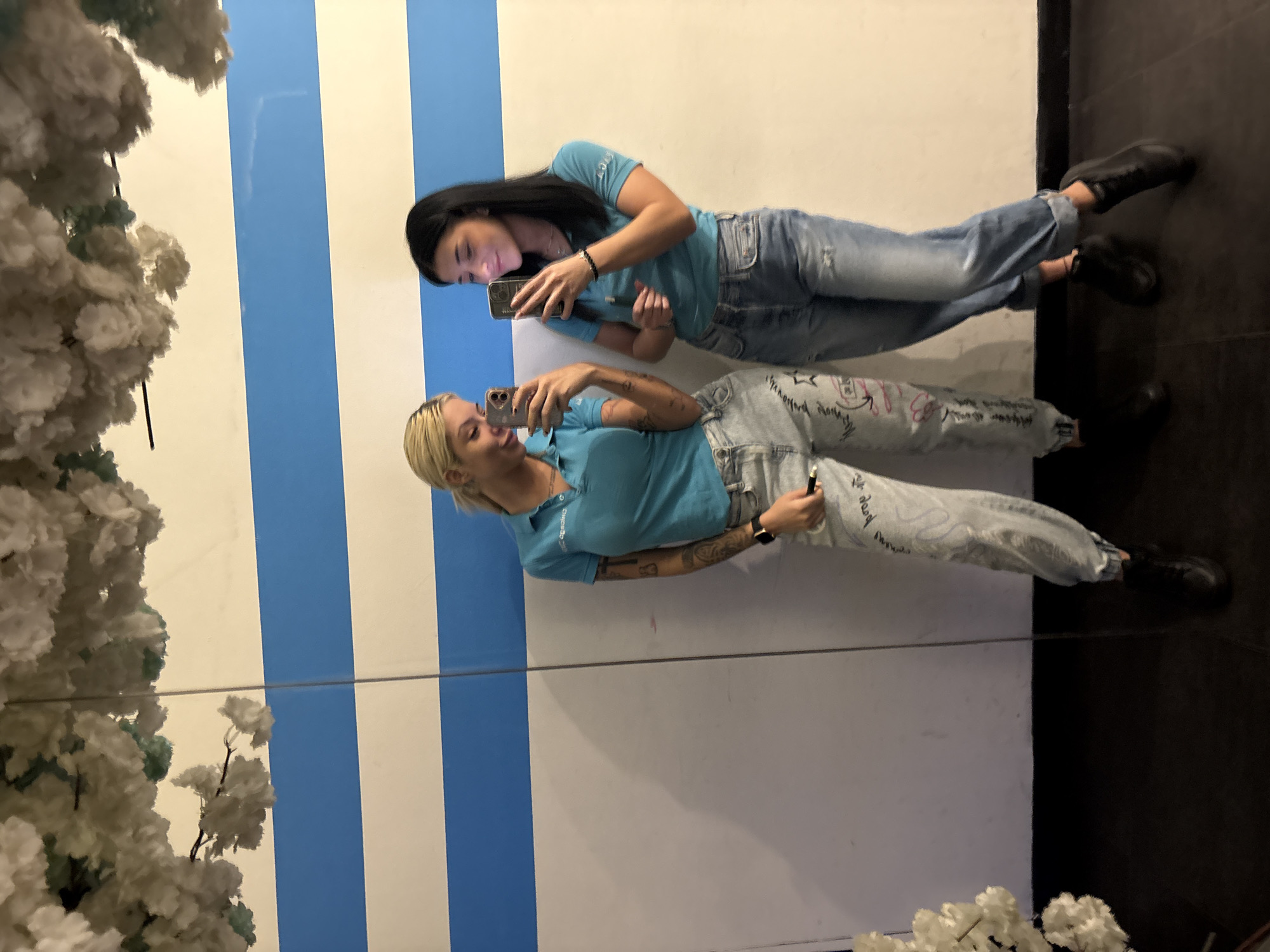 Two women stand in front of a mirror, each holding a smartphone. The woman to the left has blonde hair and is wearing a light blue t-shirt, patterned jeans, and black shoes. The woman to the right has black hair and is wearing a light blue t-shirt, jeans, and black shoes. They appear to be taking photos of themselves or each other in the mirror. The background consists of alternating blue and white vertical stripes. A floral arrangement is visible on the left side of the frame. Based on available information, the location is in the city of Plovdiv, Bulgaria.