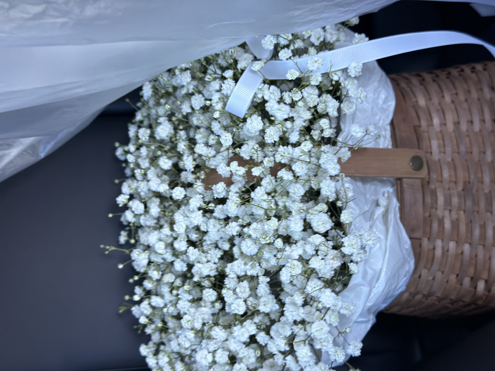 A close-up shot reveals a bouquet of baby's breath flowers nestled in a wicker basket. The flowers are delicate, white, and densely packed, with a white ribbon accenting the arrangement. A clear plastic wrap partially covers the flowers. The basket appears to have a wooden handle with a small, circular fastener. The background is out of focus, showing a dark surface to the left of the arrangement. No people are visible. There is no indication this image was taken in Chaikovsky, Russia.