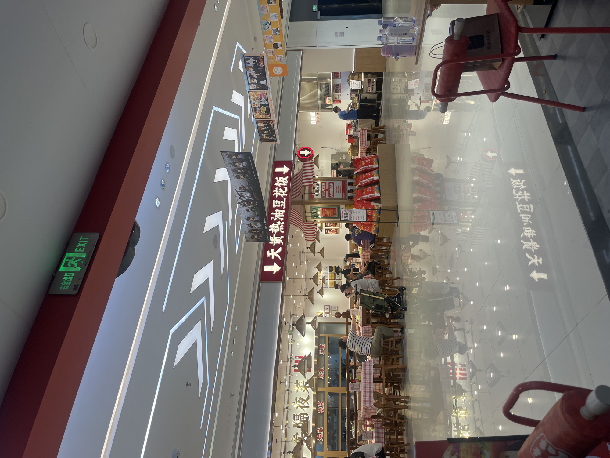 This detailed description pertains to an indoor food court or restaurant area situated within a commercial complex in Yuzhong District, China.
The scene is dominated by a brightly lit, spacious area with a highly reflective, polished light-colored tiled floor that mirrors overhead lighting and surrounding structures. The ceiling is white with distinctive angular and linear fluorescent light strips forming geometric patterns, interspersed with circular recessed lights. A prominent red structural beam runs across the ceiling, from which an illuminated green "安全出口 EXIT" sign is mounted on the left side.
In the mid-ground, a food establishment named "天贯热油豆花饭" (Tianguan Hot Oil Tofu Rice) is centrally positioned, indicated by a large red sign with white characters and a downward arrow. To its left is another restaurant, "幸福夜菜" (Happiness Night Cuisine), identified by gold characters on a white backdrop, with smaller signs below indicating "炖菜" (stewed dishes), "炒菜" (stir-fried dishes), "蒸菜" (steamed dishes), and "豆花" (tofu pudding).
The dining area features numerous square wooden tables covered with red and white checkered tablecloths, surrounded by simple wooden stools. Approximately ten to fifteen individuals are present, predominantly seated at these tables, engaged in dining and conversation. One woman is seated alongside a dark-colored baby stroller.
On the right side of "天贯热油豆花饭," a "自助区" (self-service area) is visible, where a man in a blue shirt stands behind a counter, appearing to be involved in food preparation or service. In front of the "天贯热油豆花饭" counter, a display stand holds multiple red bags.
Hanging from the ceiling are several rectangular banners adorned with anime-style characters and illustrations.
In the foreground on the right, a solitary red metal chair stands, with a small brown paper bag resting on its seat. On a nearby small wooden table, a pink thermos bottle and several clear plastic water bottles are placed. The bottom left foreground shows a partial view of a red object with a handle, possibly a child's toy or part of a piece of furniture. Reflections of overhead lights and floor markings, including upward arrows and Chinese characters, are clearly visible on the polished floor.