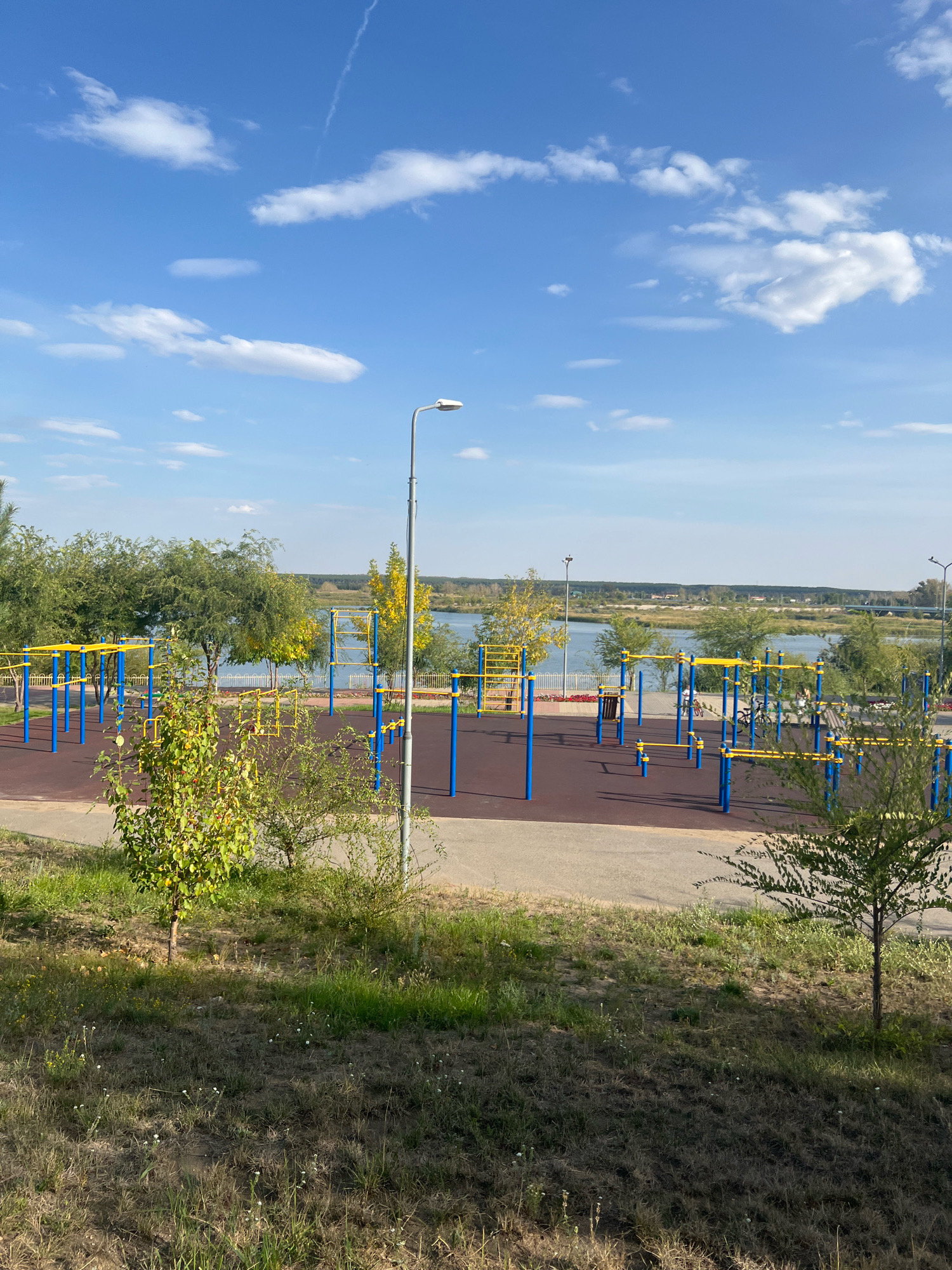 The image captures an outdoor fitness park situated beside a body of water, likely a river or lake, in Kostanay, Kazakhstan. The park features numerous pieces of exercise equipment, characterized by blue poles and yellow bars, set on a dark brown rubberized surface. The equipment includes pull-up bars, parallel bars, and other apparatus designed for bodyweight training.
The area is bordered by a pathway, and beyond that, lush green trees and grass are visible. In the background, the expanse of water stretches towards a distant treeline and a faint, possibly rural, landscape. The sky above is a clear, bright blue, punctuated by scattered white cumulus clouds, with a thin contrail from an aircraft visible. A street lamp stands prominently on the right side of the frame, near the foreground. The overall scene suggests a well-maintained public space designed for recreation and exercise, harmoniously integrated with its natural surroundings.