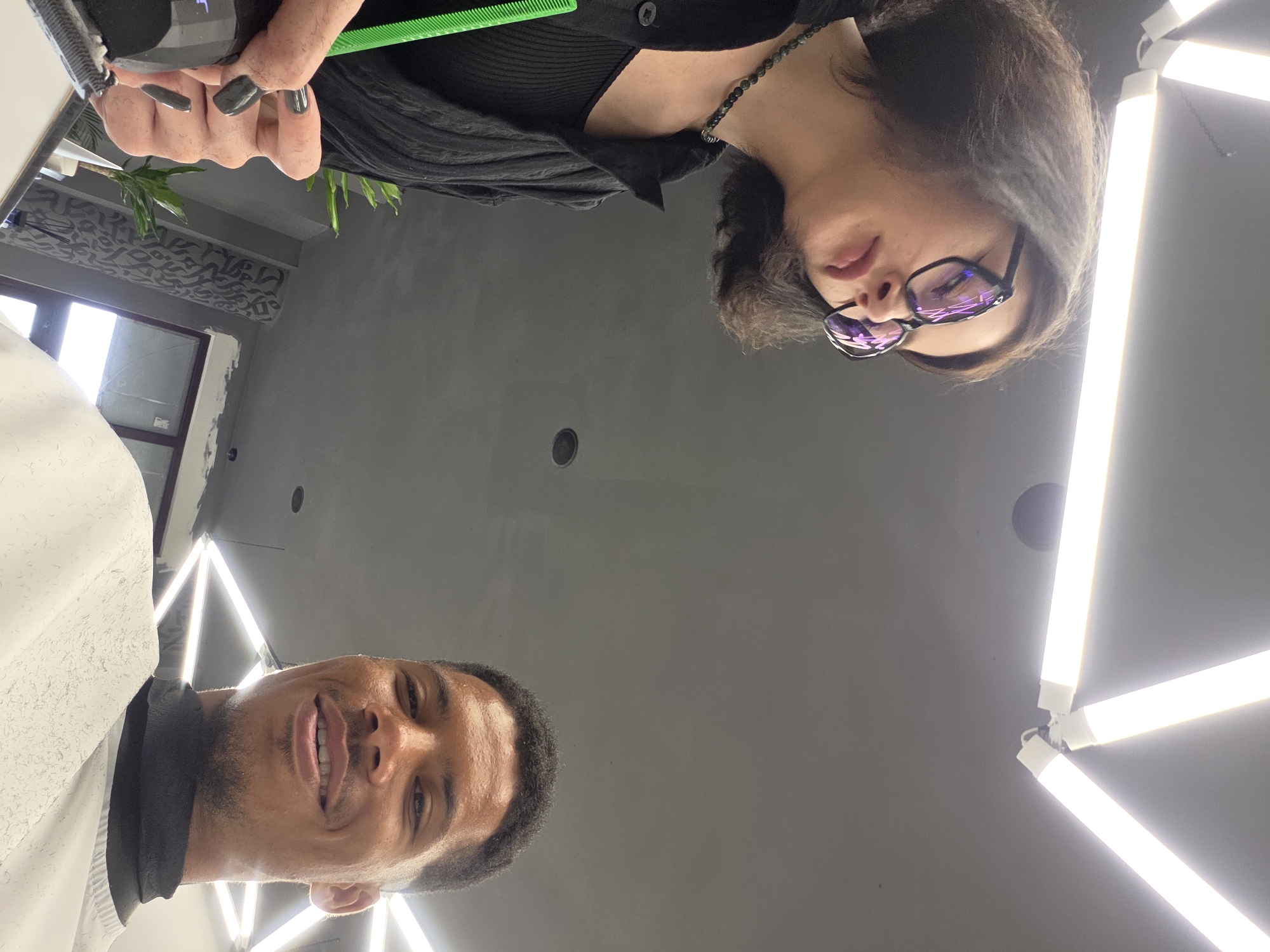 A low-angle, selfie-style shot captures two individuals in what appears to be a barbershop or salon, possibly in Varna, Bulgaria. In the foreground, a person with dark, short hair and distinctive large-framed glasses is seen from the chest up, looking down and slightly away from the camera. They are wearing a black top and a dark necklace. Their right hand, adorned with dark nail polish, is holding a bright green comb.
To the right and slightly lower in the frame, a man with short, dark hair and a close-cropped beard smiles at the camera, his face filling the lower right portion of the image. He is wearing a white garment draped over his shoulders, with a black neckline visible.
The background is dominated by a grey ceiling with a unique lighting fixture composed of several bright, linear fluorescent tubes arranged in a geometric pattern. The overall environment appears clean and modern. The presence of salon equipment, the lighting, and the action of one person holding a comb suggest a grooming service is in progress.