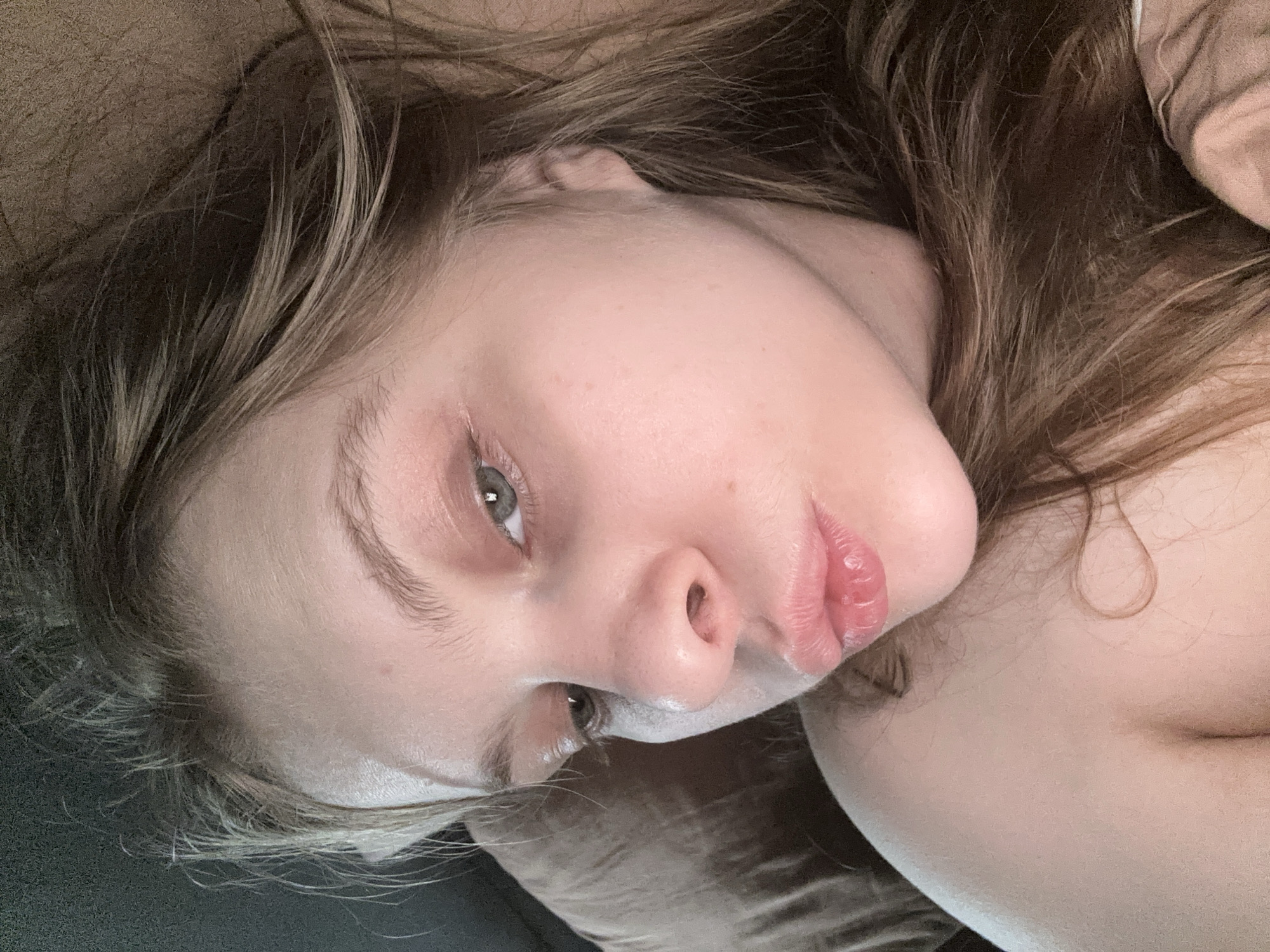 The image displays a close-up, angled portrait of a female individual lying down. Her head is positioned on a light brown textile surface, consistent with bedding or upholstery. She has fair skin, light brown hair, and light-colored irises, appearing green-grey. Her right eye is clearly visible, exhibiting some redness in the lower eyelid, and is directed towards the viewer/camera. Her lips are parted, revealing no teeth. The immediate background includes a dark, featureless surface to the upper left and a light brown, textured fabric surface to the lower right. The visual information in the image does not permit determination of a specific location within Moscow, Russia.