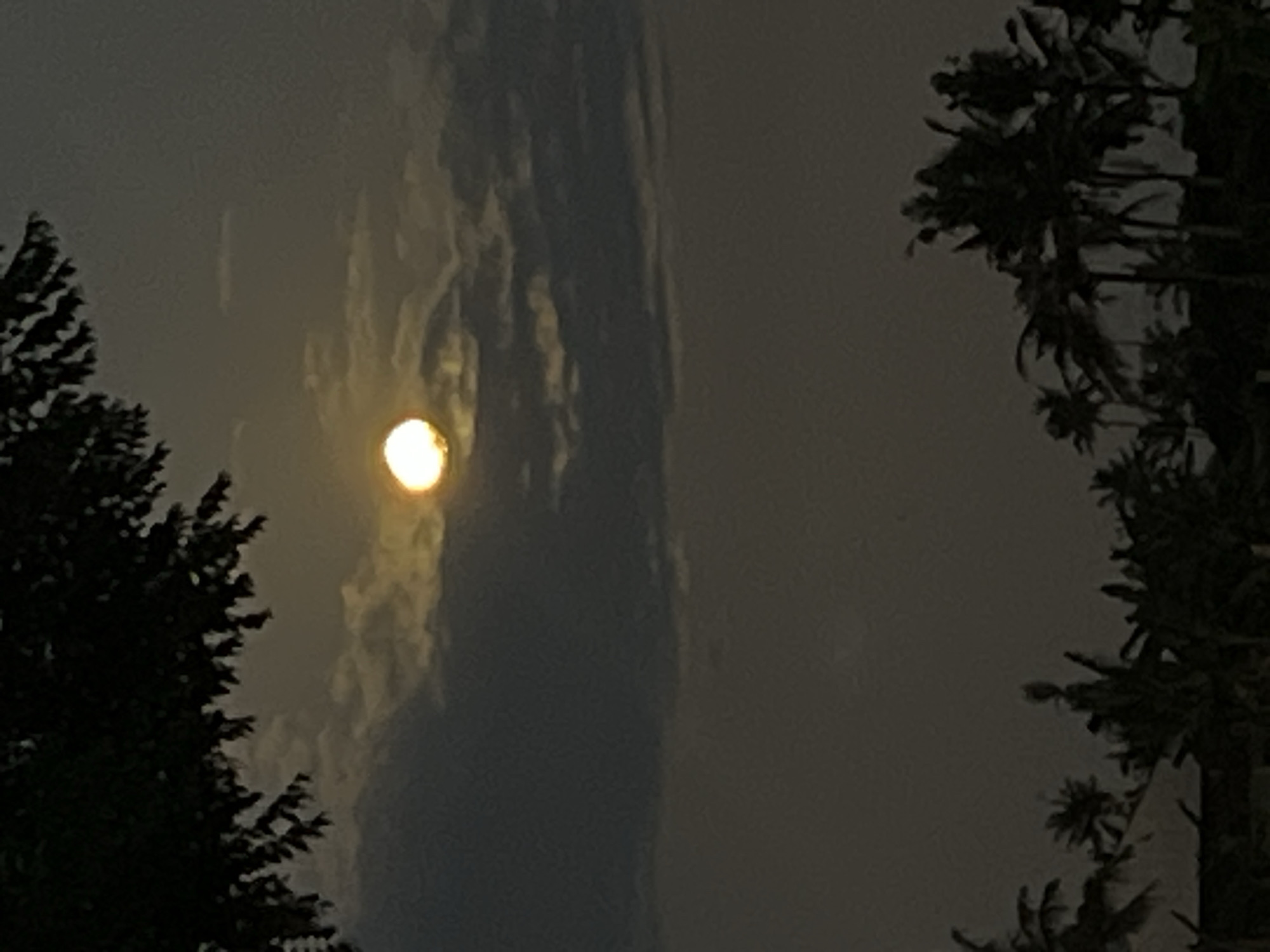 The image presents a nighttime scene, possibly from Sukawati, Indonesia, given the request context. The primary subject is a bright celestial object, likely the moon or a bright planet, partially obscured by a long, tall band of illuminated clouds. The light source casts a glow onto the clouds surrounding it. Silhouetted trees are present on both the left and right edges of the frame, their dark shapes contrasting against the lighter sky. The overall environment appears to be dark and overcast.