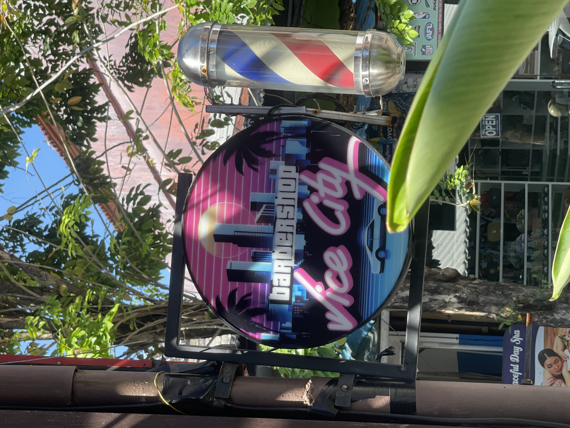 The image depicts a barbershop sign in Kuta, Indonesia. The sign is round and features a "Vice City" theme with palm trees, skyscrapers, and pink and blue hues. Above the sign is a traditional barber pole with red, white, and blue stripes. The sign is attached to a metal frame mounted on a brown structure, possibly a building exterior. A leafy plant partially obscures the view on the right side. In the background, there are other signs, foliage, and a glimpse of the sky. One sign reads "Peaceful Day Spa" and another contains the AFL and NFL logos. Legian Street No. 350 K is also listed.