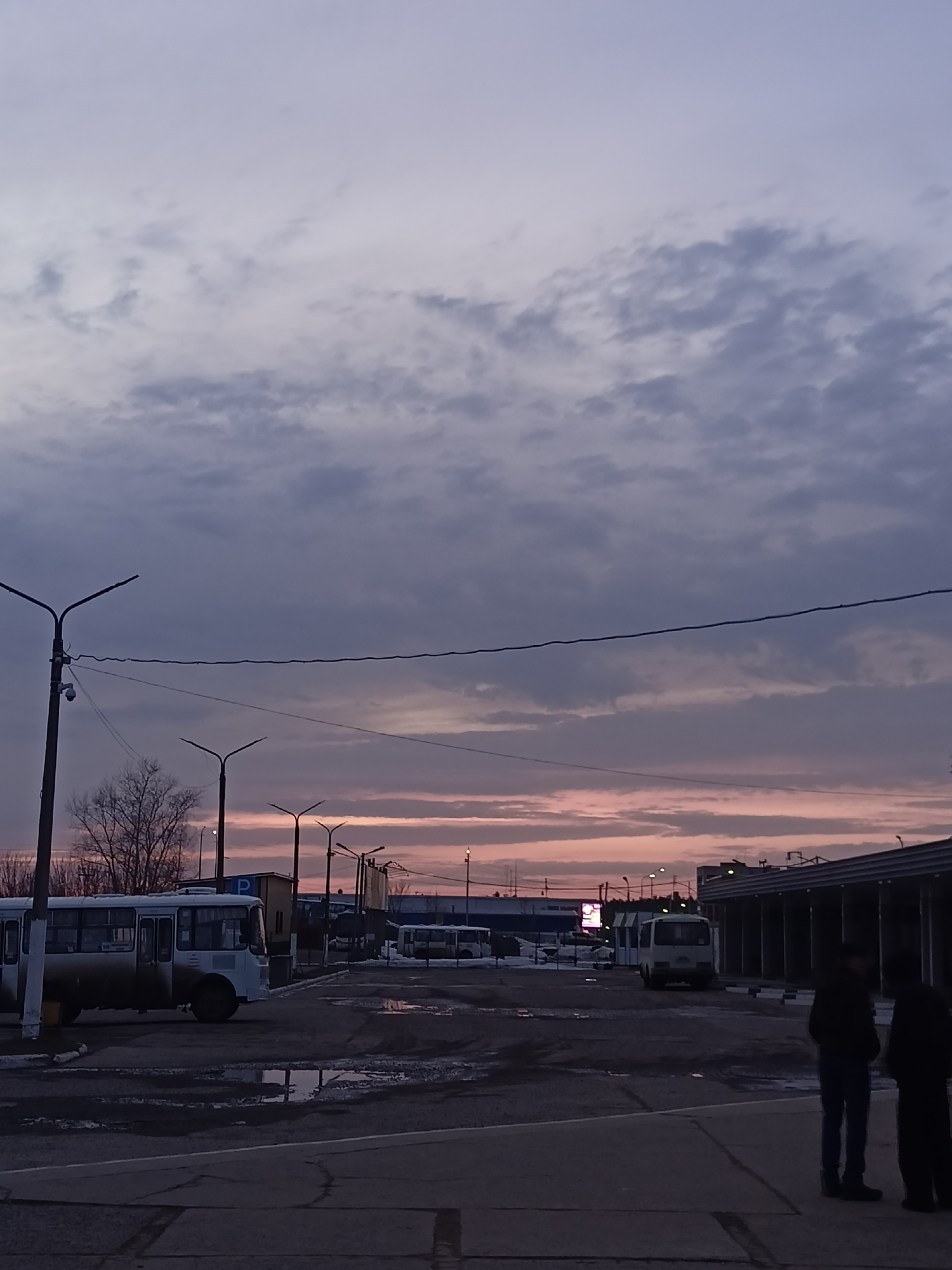 The image depicts an outdoor scene in what appears to be a bus station or transportation hub in Chaikovsky, Russia, based on the presence of shuttle vans and the general urban environment. The sky is overcast with clouds, and the light suggests it is either early morning or late evening. A pinkish hue is visible on the horizon, hinting at sunrise or sunset.
In the foreground, there is an expanse of paved area, likely a platform or parking space, which is wet in several spots, indicating recent rain or snowmelt. Patches of what appears to be residual snow or ice are visible on the ground. To the left, a light-colored shuttle van is parked; its details are somewhat obscured by the dim lighting. A few street lamps illuminate the area.
In the middle ground, several more shuttle vans are parked. In the right foreground, two dark figures are standing, presumably people waiting for transportation, and partially obscured. In the background, there are additional buildings and infrastructure elements, characteristic of an urban setting.