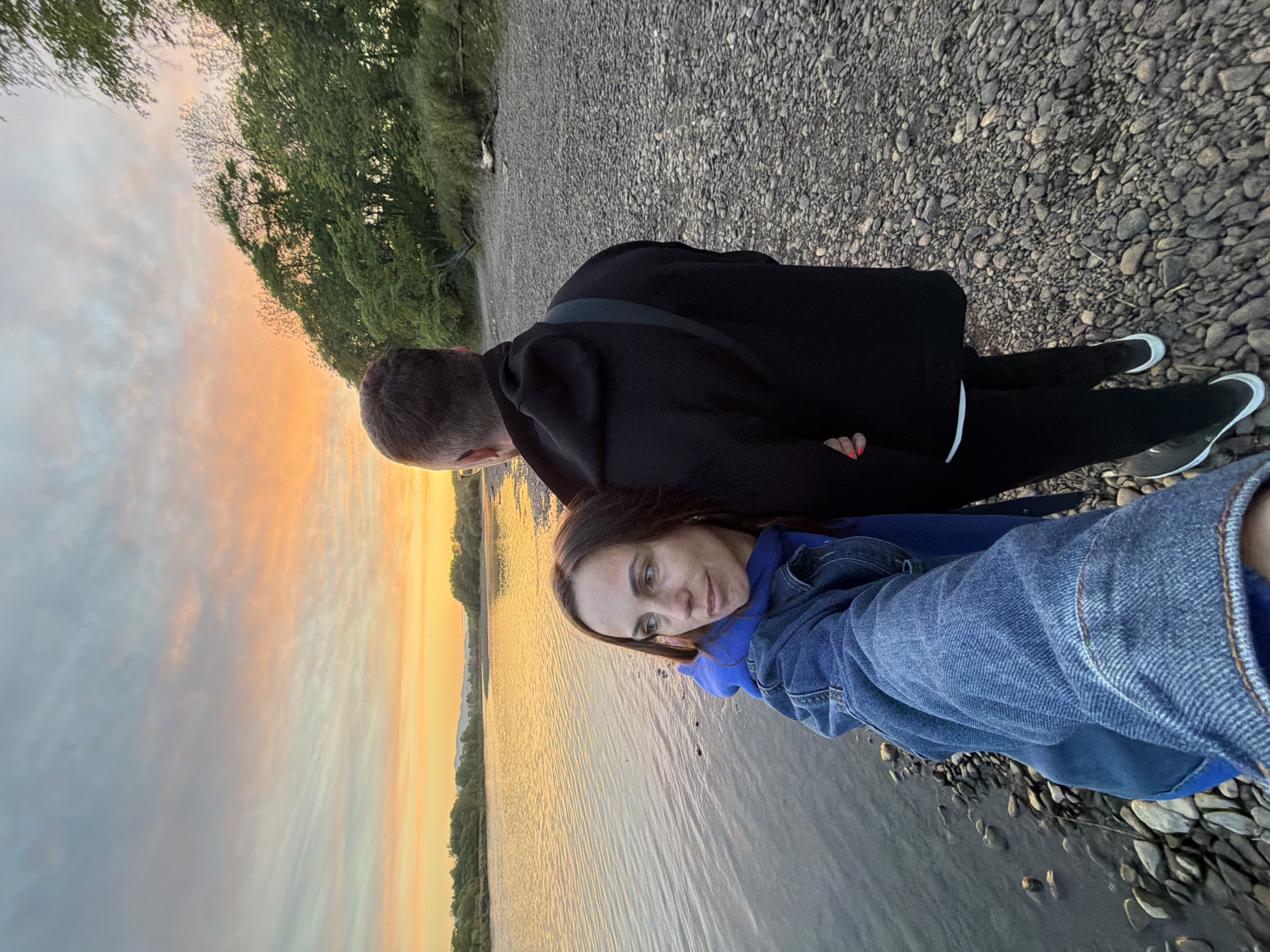 Two people are standing on a stony bank next to a body of water, potentially a river or lake, at sunset. One person is wearing a blue jacket and the other is wearing a black jacket and black pants. The background includes trees and a sky with orange and yellow hues from the setting sun. It is not possible to determine the location as Birobidzhan, Russia from the image.