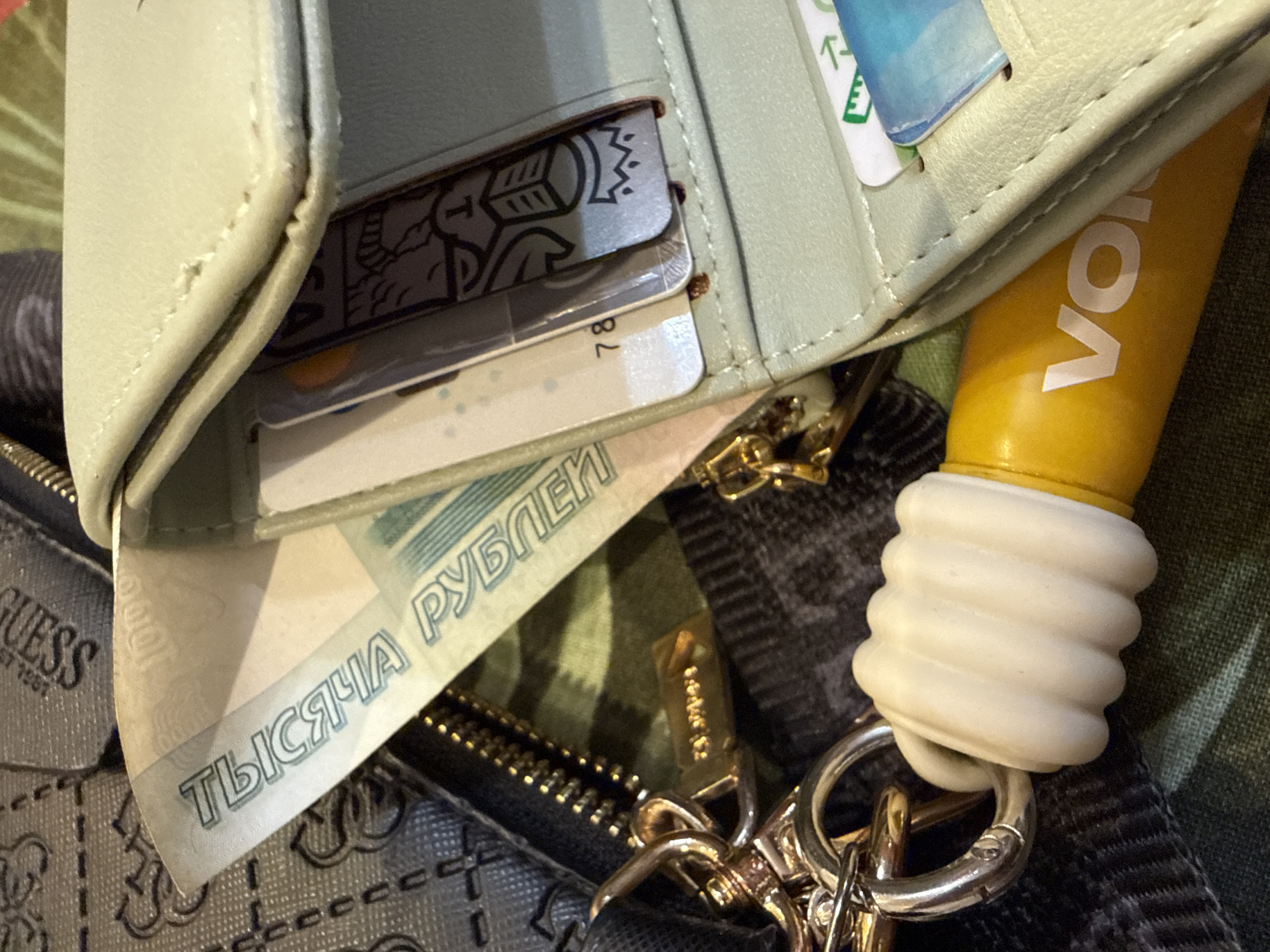 This close-up image displays an arrangement of personal belongings, likely belonging to an individual in Dmitrov, Russia. The central subject is an open, light mint green wallet made of what appears to be leather or faux leather. Inside the wallet, several cards are visible; one card partially shows the numbers "78".
A Russian 1000-ruble banknote is prominently featured, sticking out from the wallet's fold, clearly displaying the text "ТЫСЯЧА РУБЛЕЙ" (ONE THOUSAND RUBLES) in Cyrillic.
To the right, a bright yellow cylindrical object, possibly a lip balm or small tube, is visible, bearing the partial white text "VO". Below this object is a metal keychain with multiple rings, attached to a distinctive white, ribbed rubber or plastic accessory. The keychain is connected to a larger item, possibly a bag.
In the bottom left and stretching across the bottom, a grey bag or purse is partially visible, featuring a textured pattern and the brand name "GUESS" along with "EST 1981". A zipper with a small metallic pull is also seen. The background consists of a dark green, possibly camouflage-patterned fabric. The overall scene suggests an indoor setting, with no specific activity or time of day discernible.