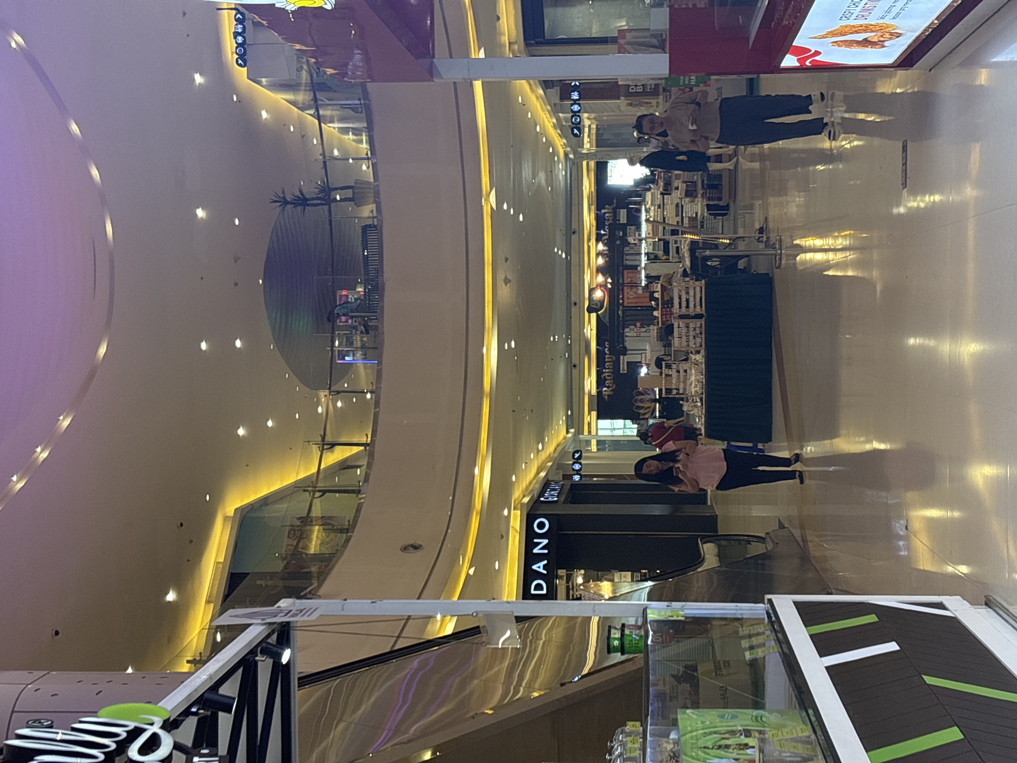 The image depicts an interior view of a brightly lit shopping mall in North Jakarta, Indonesia. The perspective is from the ground floor looking down a wide corridor with multiple levels visible above. The mall features a modern architectural design with a curved ceiling illuminated by numerous recessed lights and accentuating yellow LED strips along the edges of different levels.
On the left side of the foreground, a section of a retail display is visible, featuring a glass-fronted cabinet with shelves stocked with products. The display has a dark brown base with diagonal green and white stripes. Adjacent to it is a polished metal escalator.
In the mid-ground, several shops are visible. A prominent sign reads "DANO" in large white letters on a black background, with "GIORDA" visible next to it, suggesting clothing stores. Further down the corridor, another store sign displays "Radiance." People are seen walking in the mall. A woman in a pink shirt and black pants is walking towards the left, looking at her phone. Another person, a man in a light-colored shirt and dark pants, is walking towards the right, also looking at his phone. In the background, more shoppers are dispersed, and displays of merchandise are apparent. The floor is highly reflective, mirroring the overhead lights and architectural elements. On the upper levels, balconies and railings are visible, offering glimpses into other parts of the mall. The overall ambiance is that of a busy commercial space.