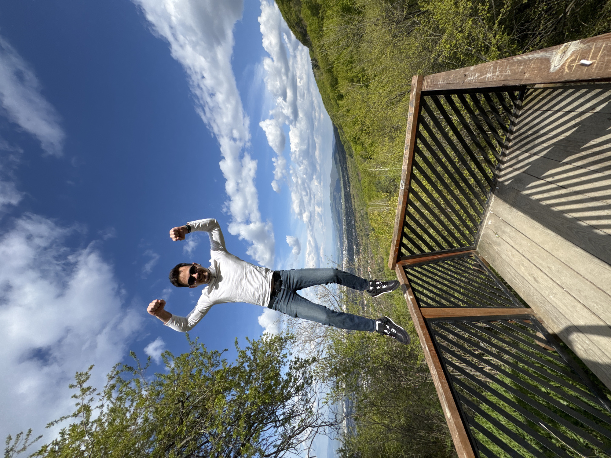 A man in a white long-sleeve shirt, jeans, and black shoes is captured mid-air in a playful pose with his fists clenched above his head, suggesting he is "flying." He wears sunglasses.
The photo is taken from a low angle, with the sky and clouds forming the background above the man. Below him, there's a view of a city, a forested hill, and some trees close to the camera.
To the right of the man, there's part of a wooden structure, possibly a lookout point or deck, with a railing featuring a pattern of parallel bars.
The location is the Kopitoto lookout point on Vitosha mountain in Sofia, Bulgaria.