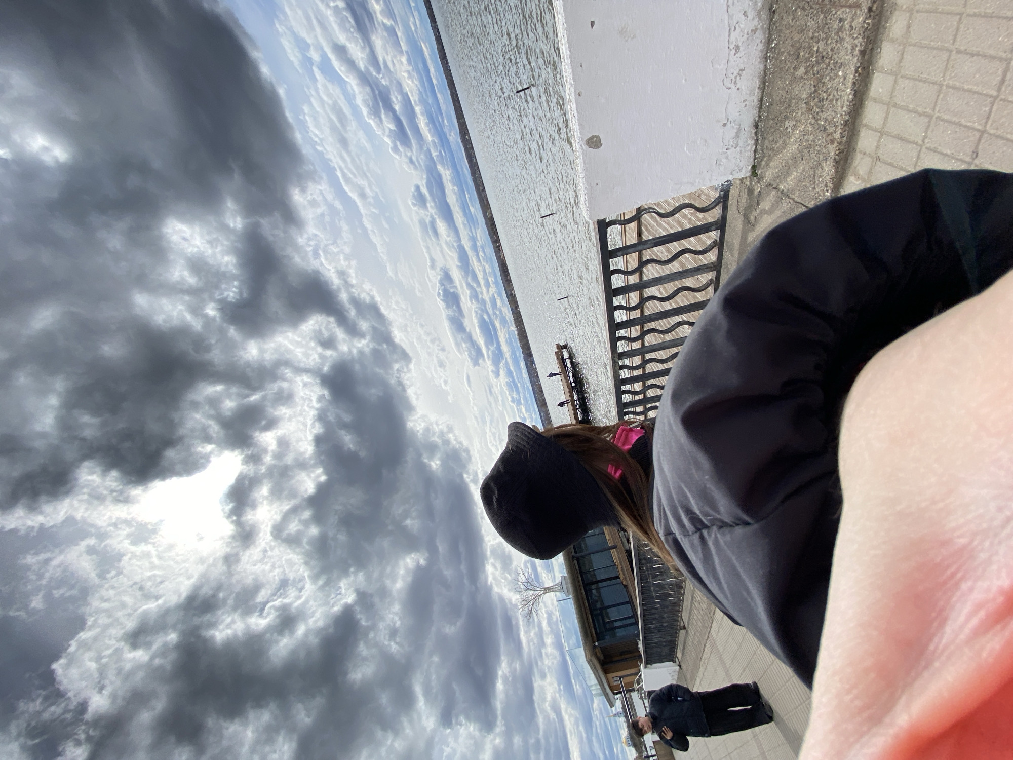 The image captures a first-person perspective of a cloudy day by a large body of water, likely in Votkinsk, Russia. In the immediate foreground, to the right, a fair-skinned arm or hand is visible, suggesting the camera is held by this person. To the left, the back of another person's head and shoulder are prominent. This individual wears a black beanie or cap, a black puffer jacket, and has brown hair with a pink accessory visible. They are looking out over the water.
A black metal railing with wavy horizontal bars stretches across the midground, separating the viewer from the water. To the right of the person in the black jacket, a white concrete wall or embankment shows signs of wear and peeling paint. The ground beneath appears to be paved with light-colored tiles.
The background features a calm, vast expanse of water, reflecting the overcast sky. On the distant shoreline, a modern-looking building with large glass windows and a unique, bare, sculptural tree on its roof is visible. Another person in a dark jacket stands on a paved area near this building.
The sky is heavily covered with dramatic, grey clouds, but patches of bright light and direct sunlight break through, indicating a day with dynamic weather. The light suggests it is daytime, possibly late morning or afternoon. There is no visible text in the image.
