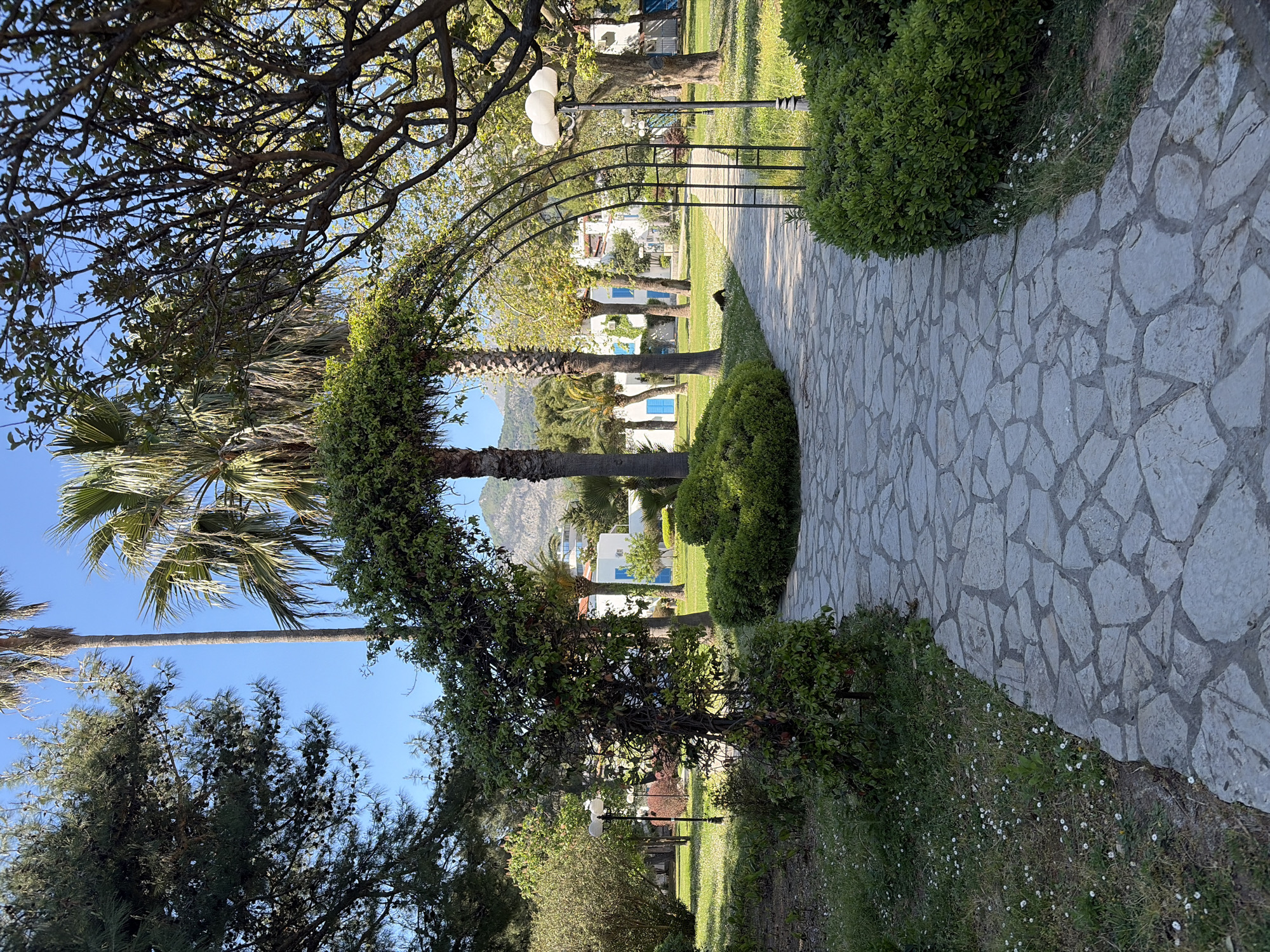 The image shows a stone-paved pathway leading through a garden or park area. The path is bordered by green bushes and trees. A metal arch covered in foliage spans the pathway. In the distance, white buildings with blue window frames are visible, along with palm trees. The setting is likely a resort or landscaped area within the city of Budva, Montenegro.