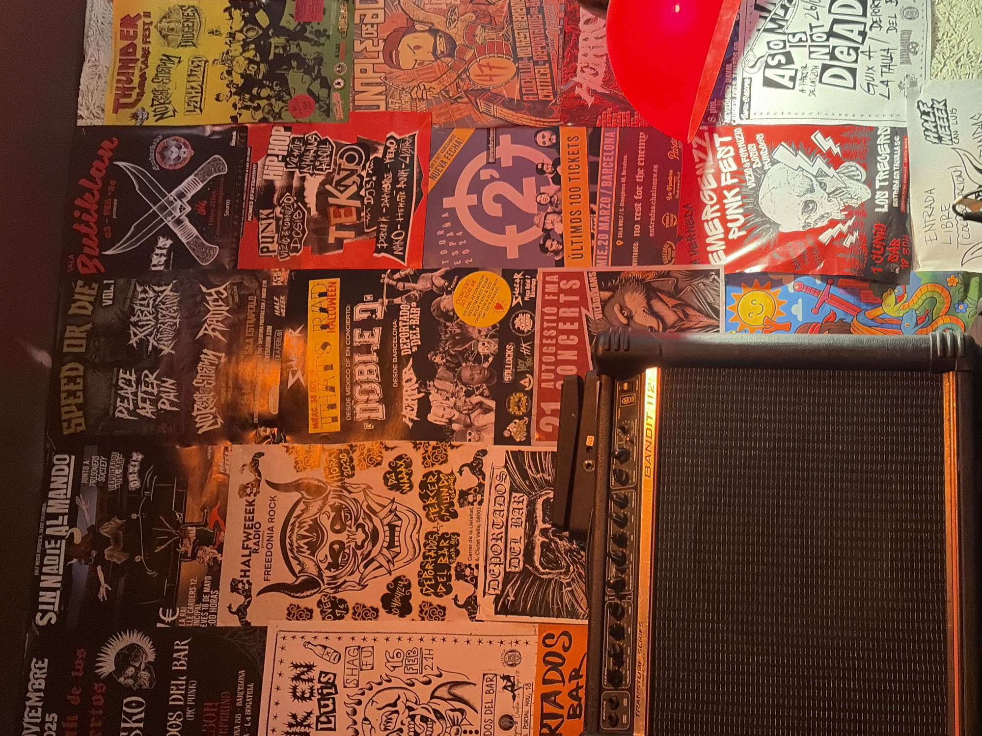 The image shows a wall covered with numerous concert posters and flyers, with a guitar amplifier in the foreground. The posters advertise various music events, predominantly in the punk and rock genres, with dates ranging from November 2025 to March 2025. Some posters feature band names like "SIN NADIE ALMANDO," "SPEED OR DIE," "BUTIKLAN," and "THUNDER." Location details on some posters include "BARCELONA" and specific venues like "SALA ESTRAPERLO" and "SALA WOLF." The guitar amplifier is a "TRANSTUBE SERIES BANDIT 112." The environment appears to be dimly lit, possibly a music venue or a backstage area. The specific location in Sant Adrià de Besòs, Spain, is not explicitly stated on the visible posters.