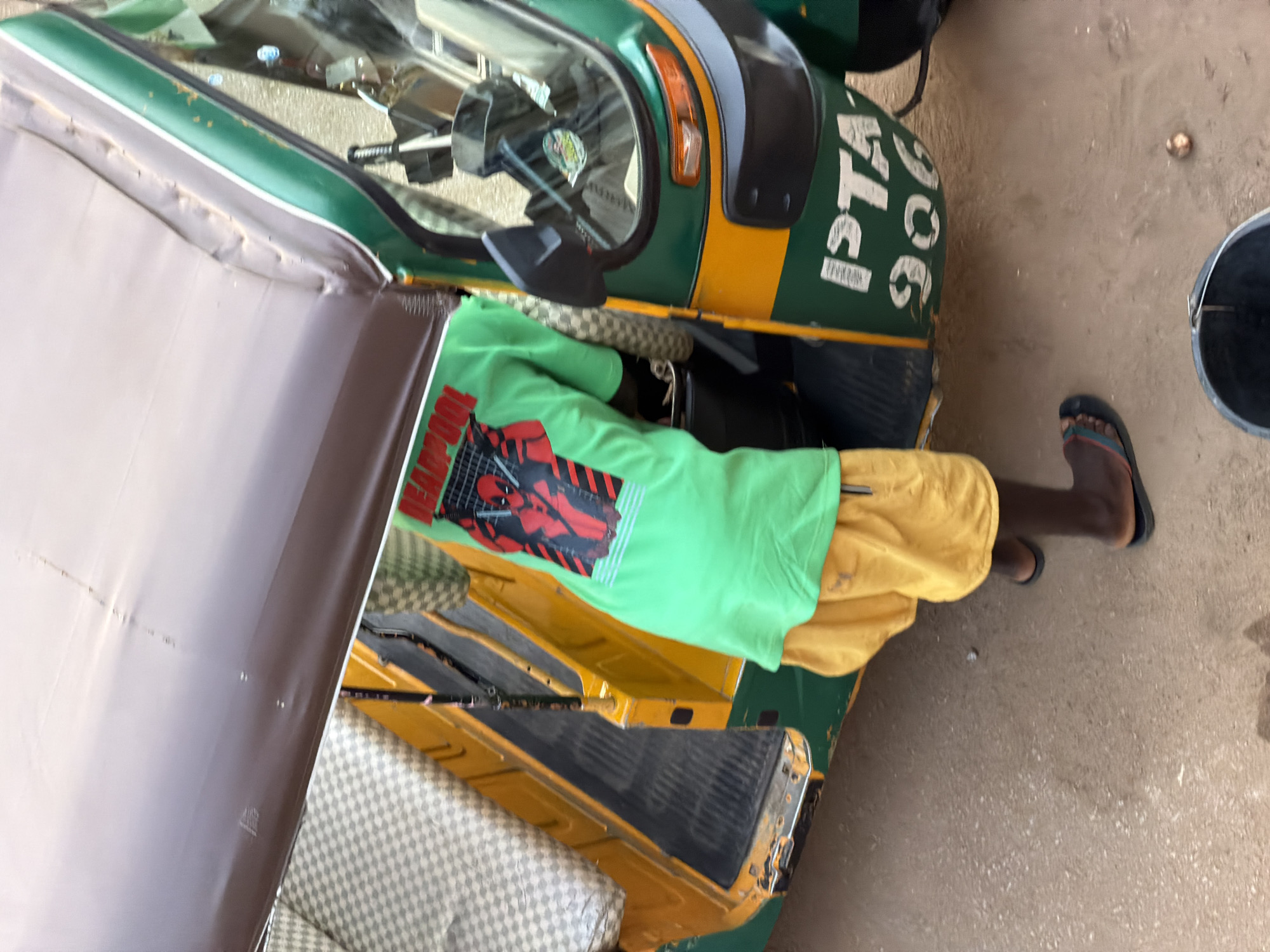In Bukuru, Nigeria, a person wearing a green t-shirt with a red and black graphic and yellow shorts stands next to a green and yellow auto-rickshaw. The person is bent over, appearing to interact with a black bucket placed inside the open door of the auto-rickshaw. The auto-rickshaw has a brown canopy and a checkered pattern on the seats. The vehicle has "PTA 906" stenciled in white on its side. The ground is a dusty, reddish-brown surface.